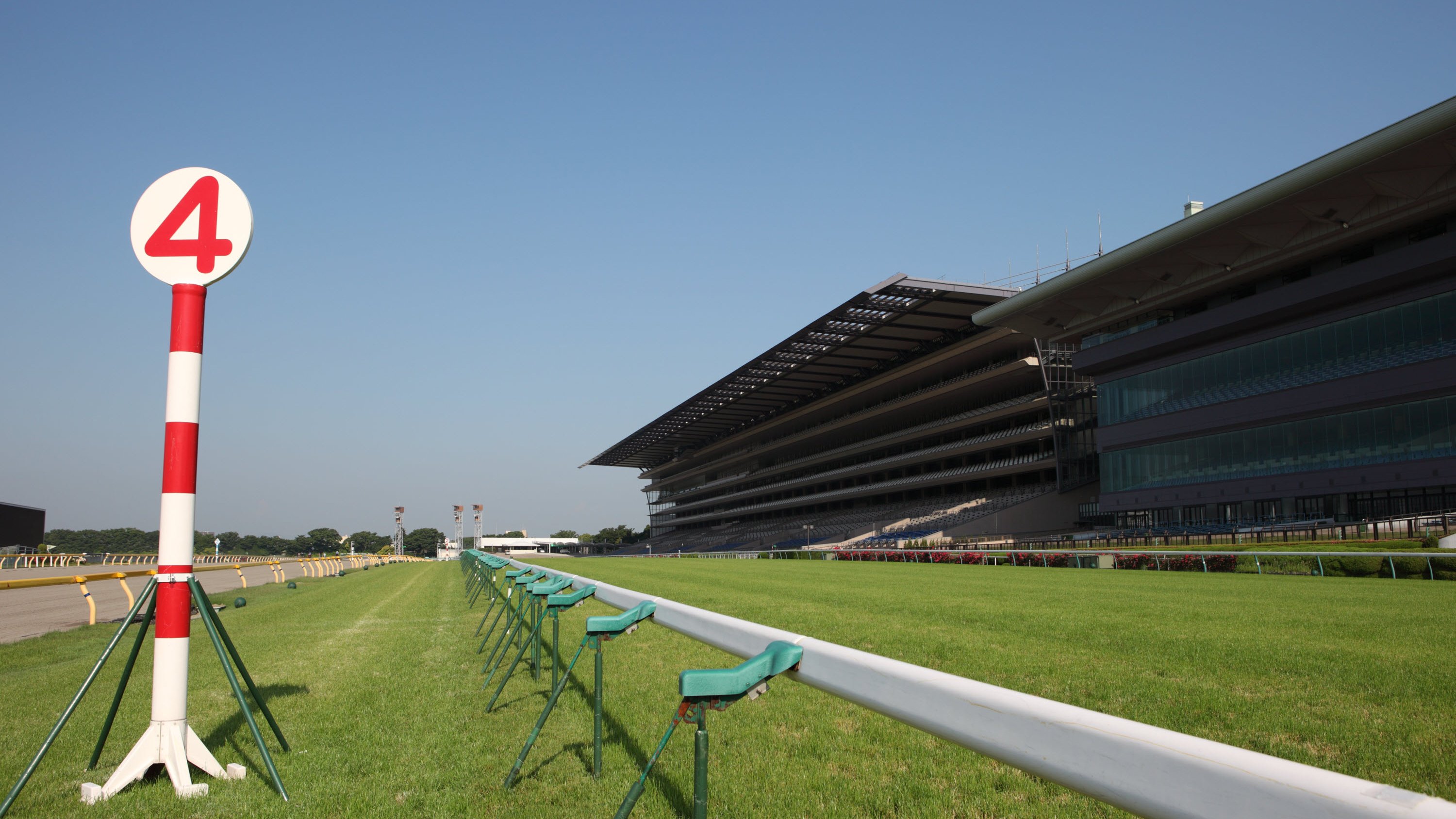 A horse racing track with a red and white pole marked '4' and a grandstand in the background under a clear blue sky.