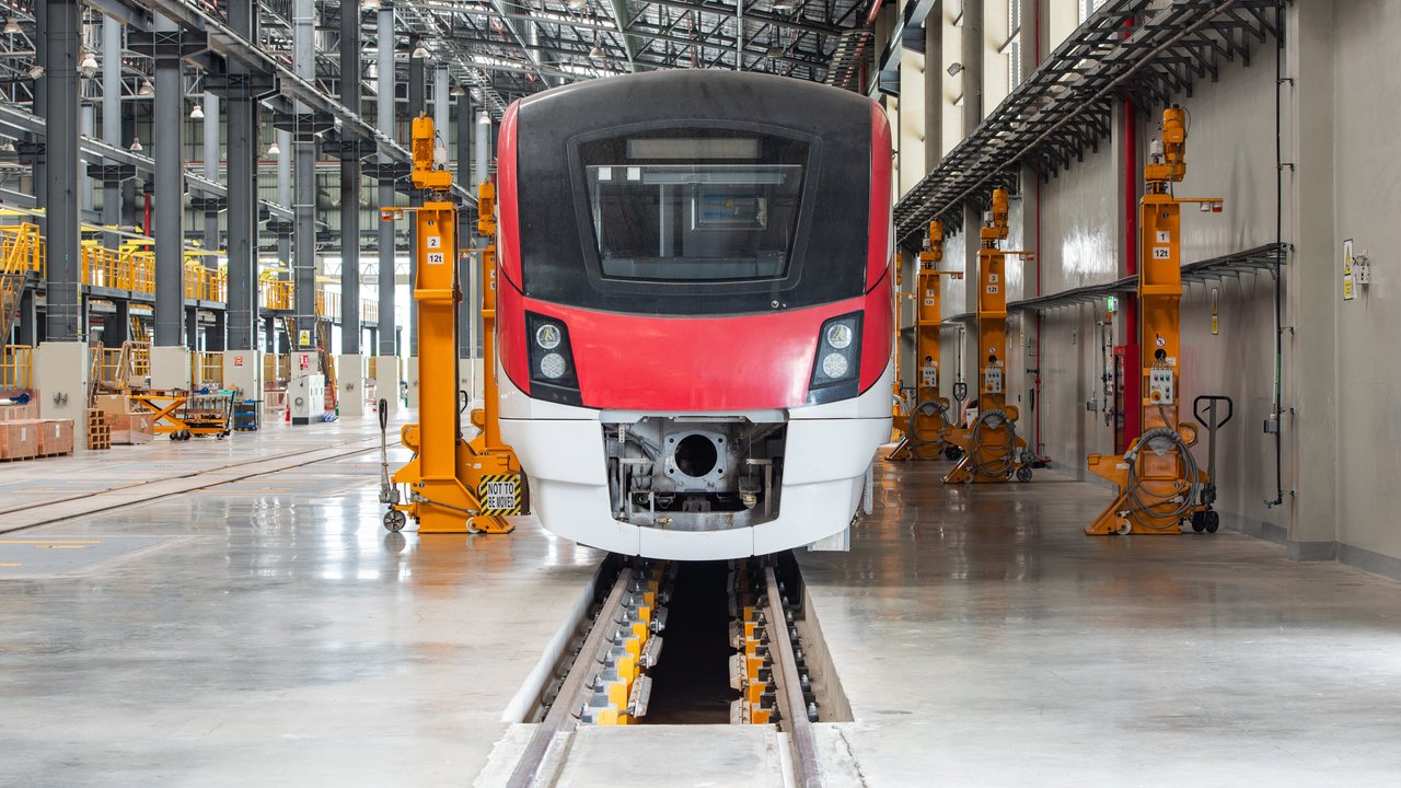 A red and white train front is positioned on tracks inside a spacious maintenance facility with yellow cranes.