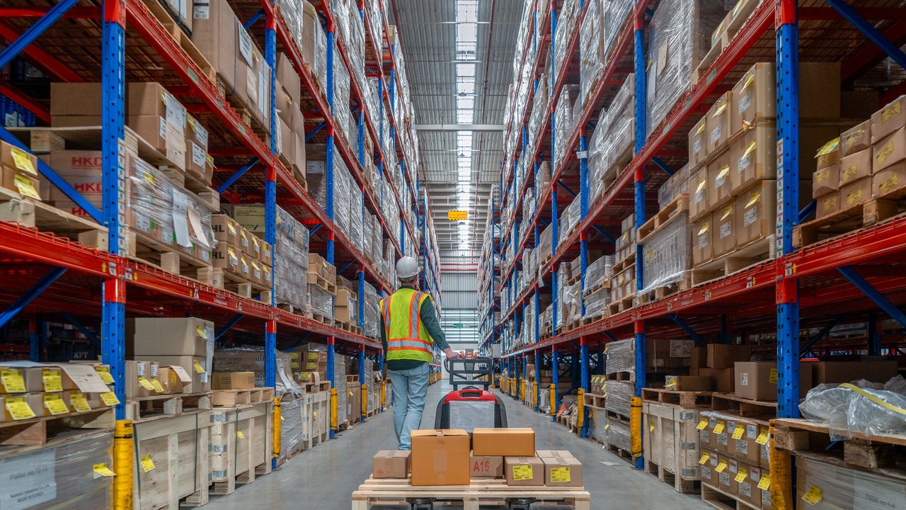 A worker pushes a pallet jack loaded with boxes in a large warehouse aisle lined with shelves of packaged goods.