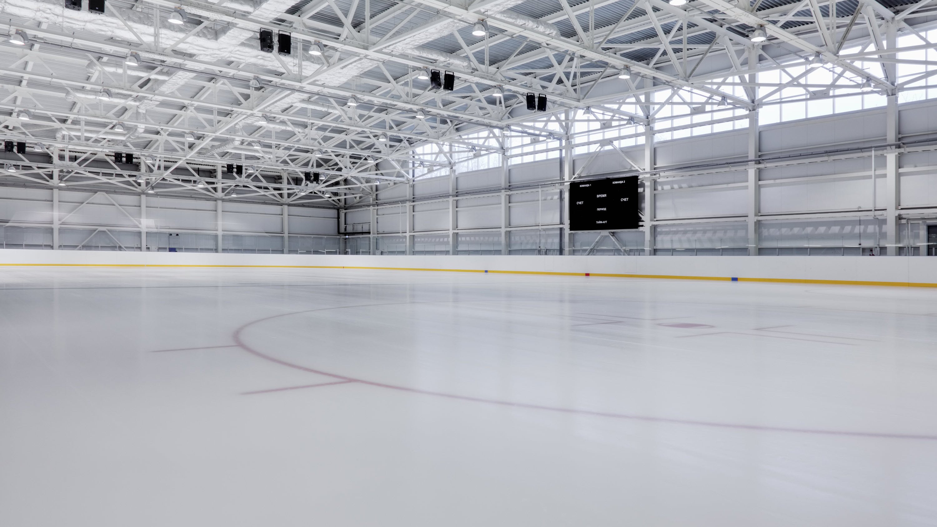 An empty ice rink with a white surface, yellow boards, and a scoreboard in a well-lit arena.