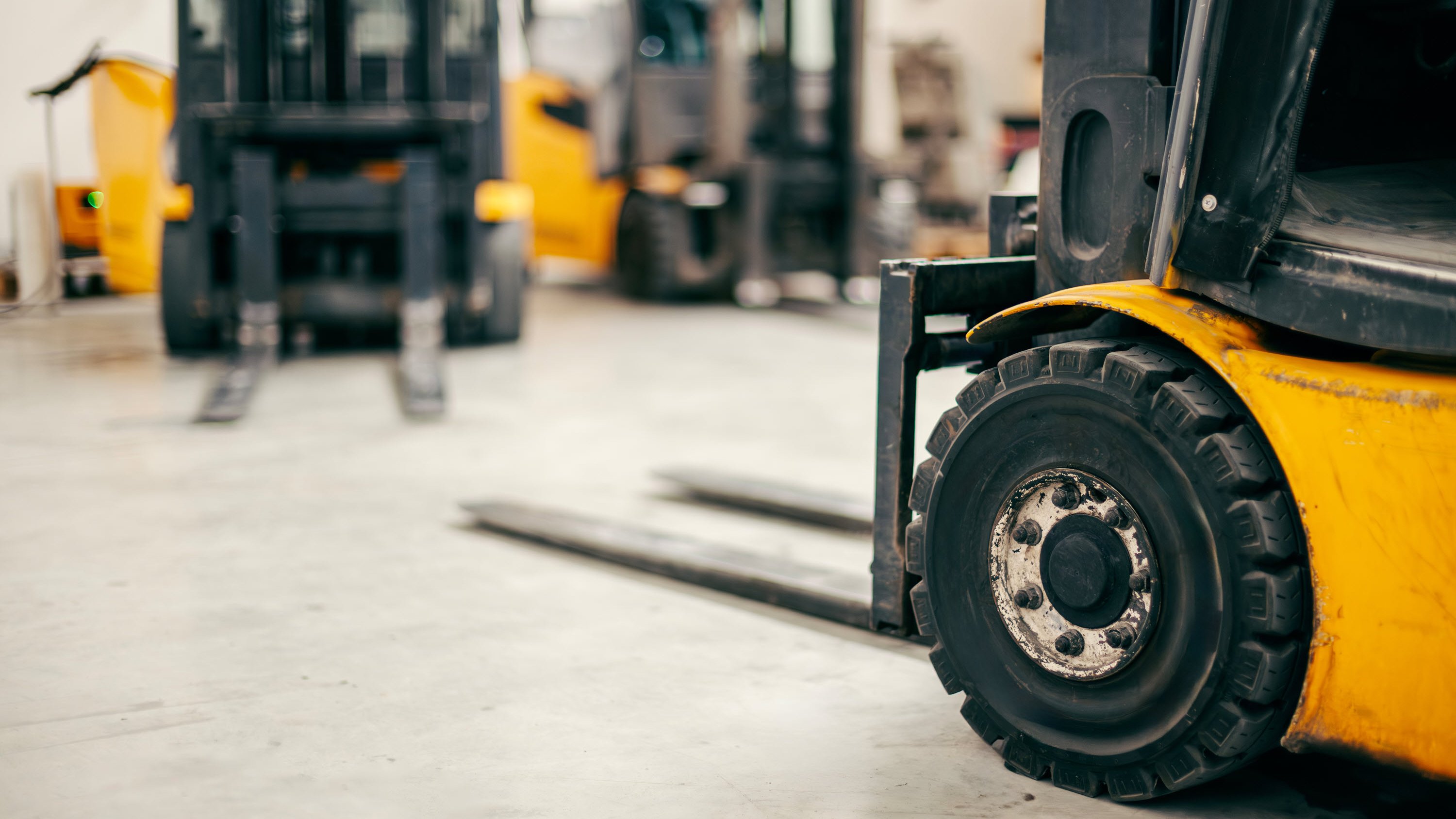 Close-up of a yellow forklift tire with blurred forklifts in the background on a concrete floor.