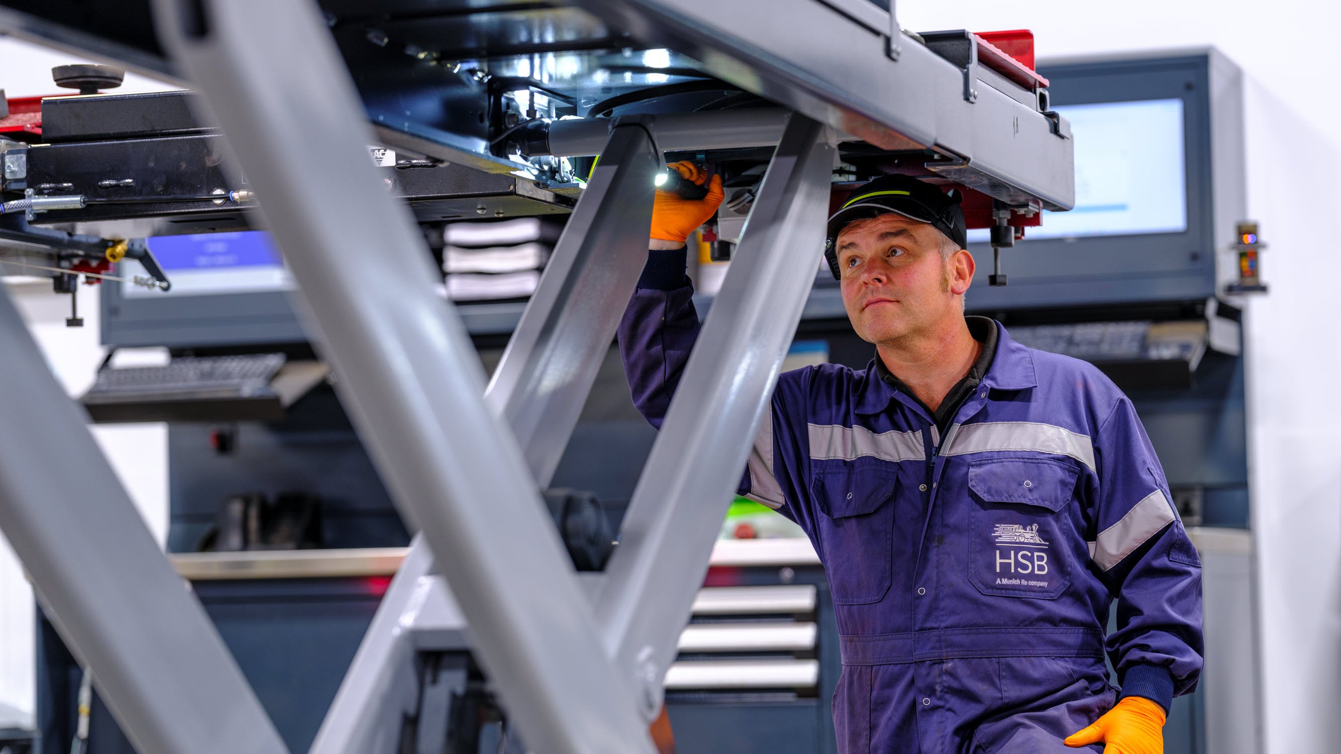 A worker in a blue jumpsuit and orange gloves operates machinery in a workshop with equipment in the background.