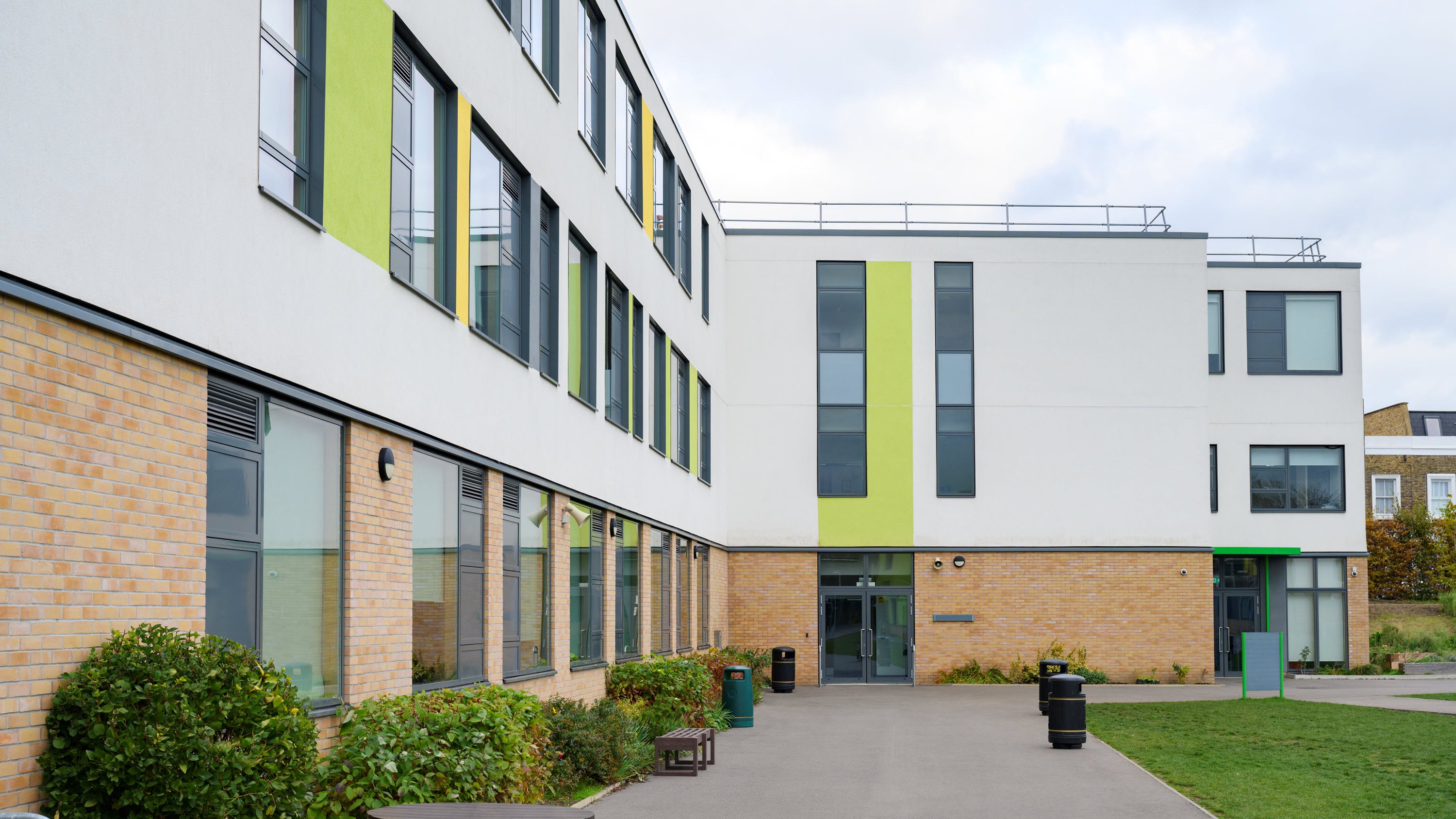 Modern building with large windows, green accents, and a paved area with benches and shrubs.