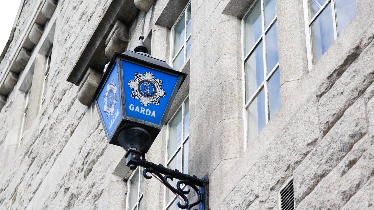 A blue Garda sign hangs from a black bracket on a stone building, with windows visible in the background.