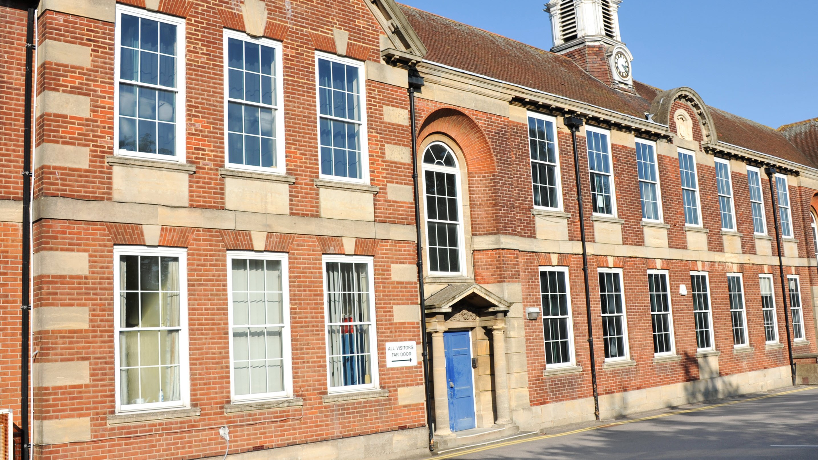 A brick school building with large windows and a blue door, featuring a clock tower on the roof.