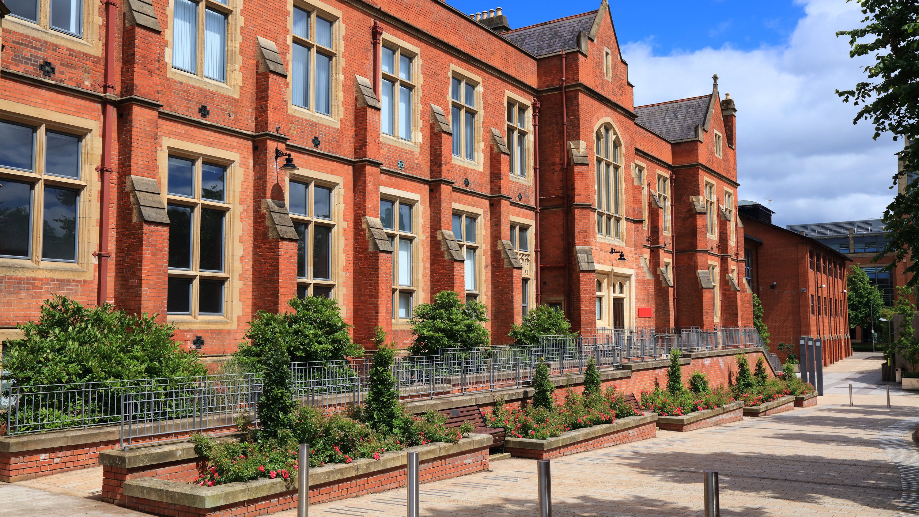 A red brick building with large windows, surrounded by greenery and a paved walkway under a blue sky.