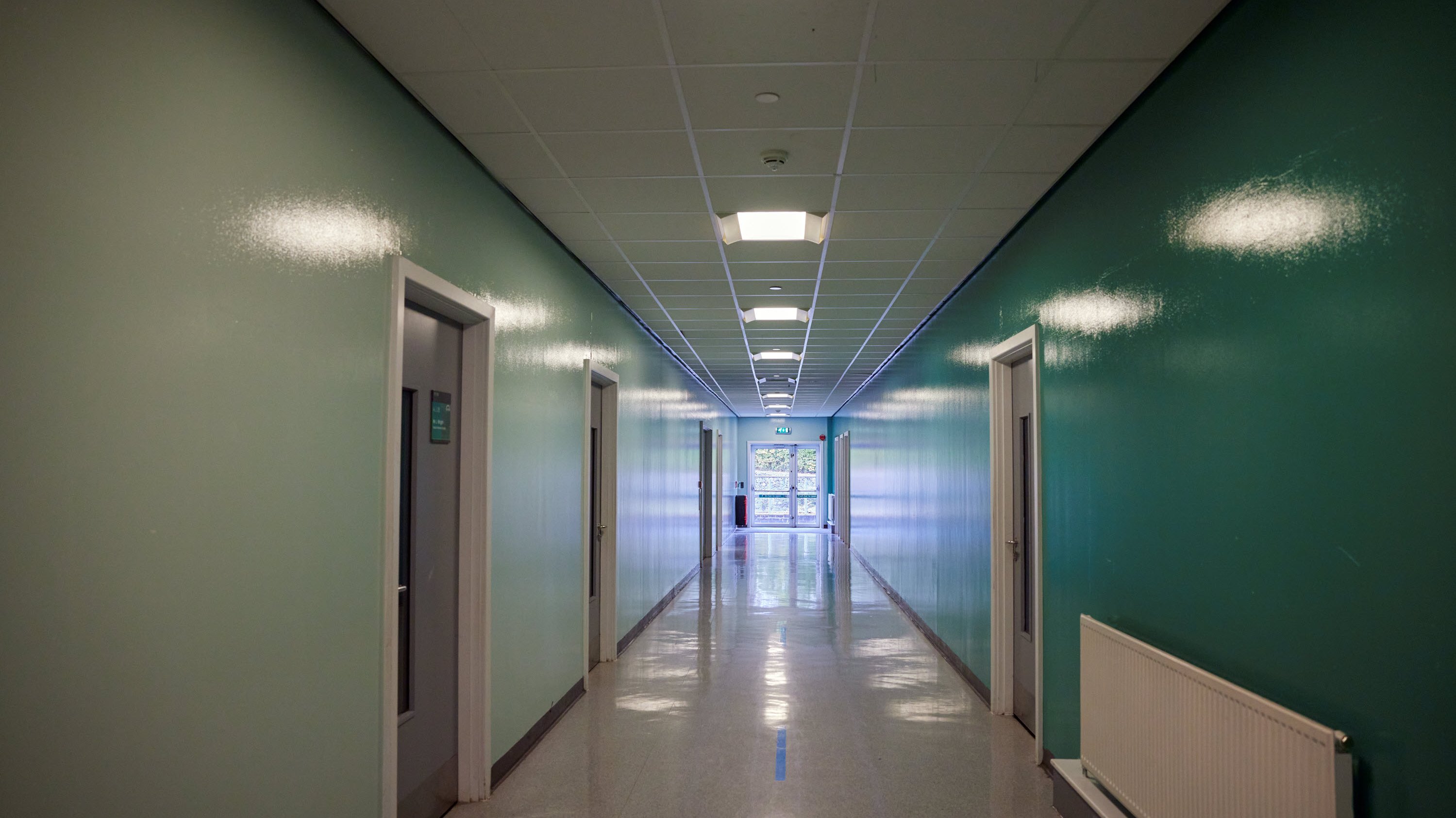 A long, brightly lit corridor with green walls and doors on either side, leading to a glass door at the end.