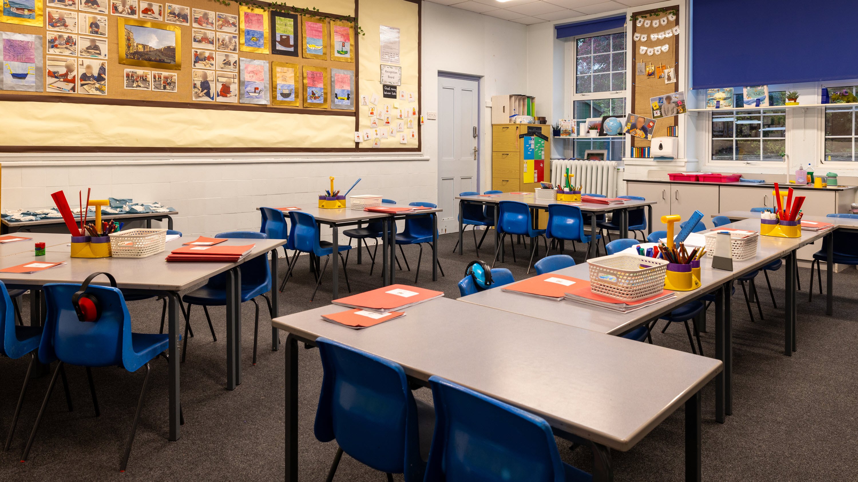 A classroom with blue chairs, tables, art supplies, and colorful student artwork displayed on the walls.
