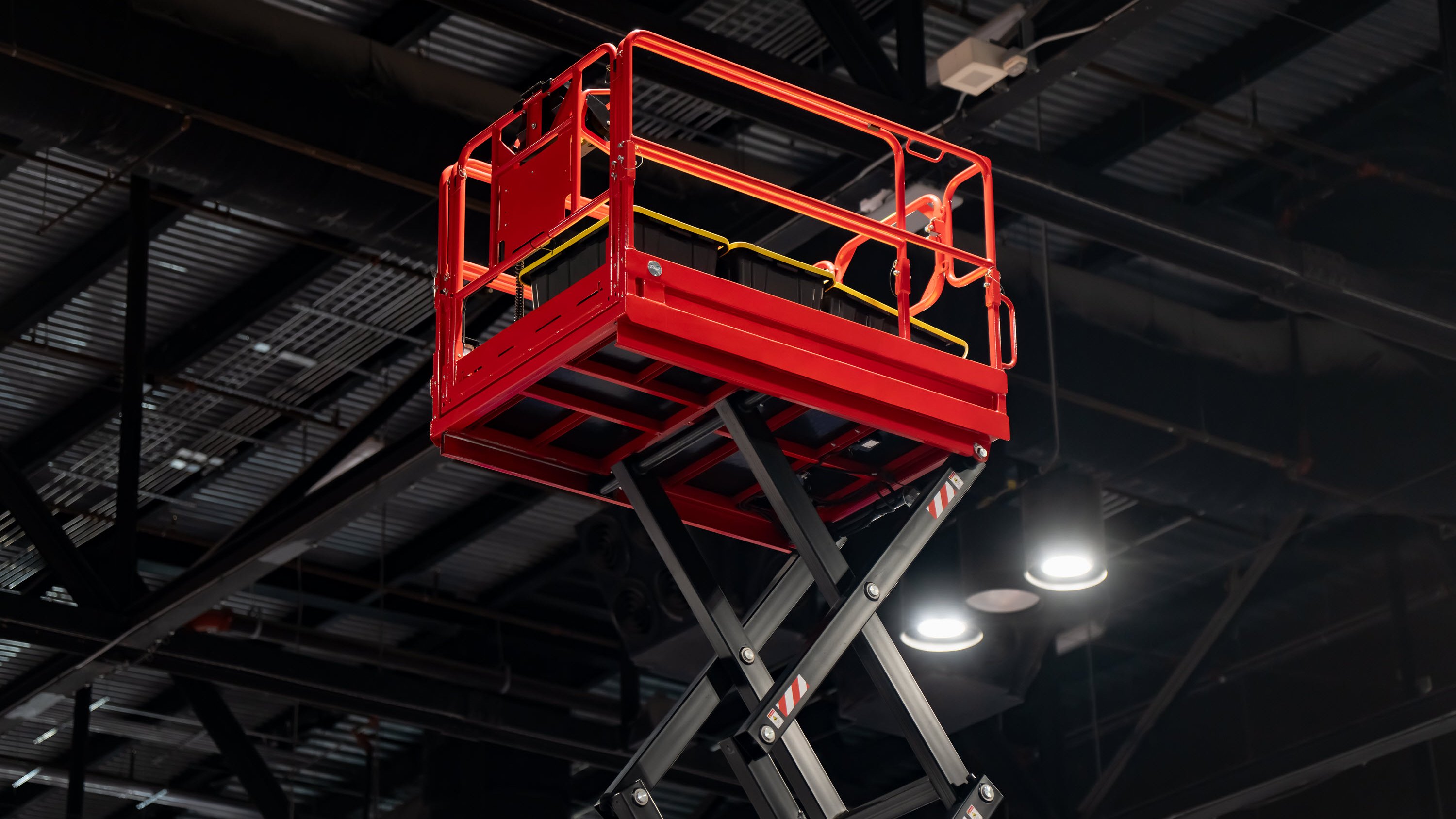 A red scissor lift platform is elevated, with a black base and safety railing, set against a dark industrial ceiling.