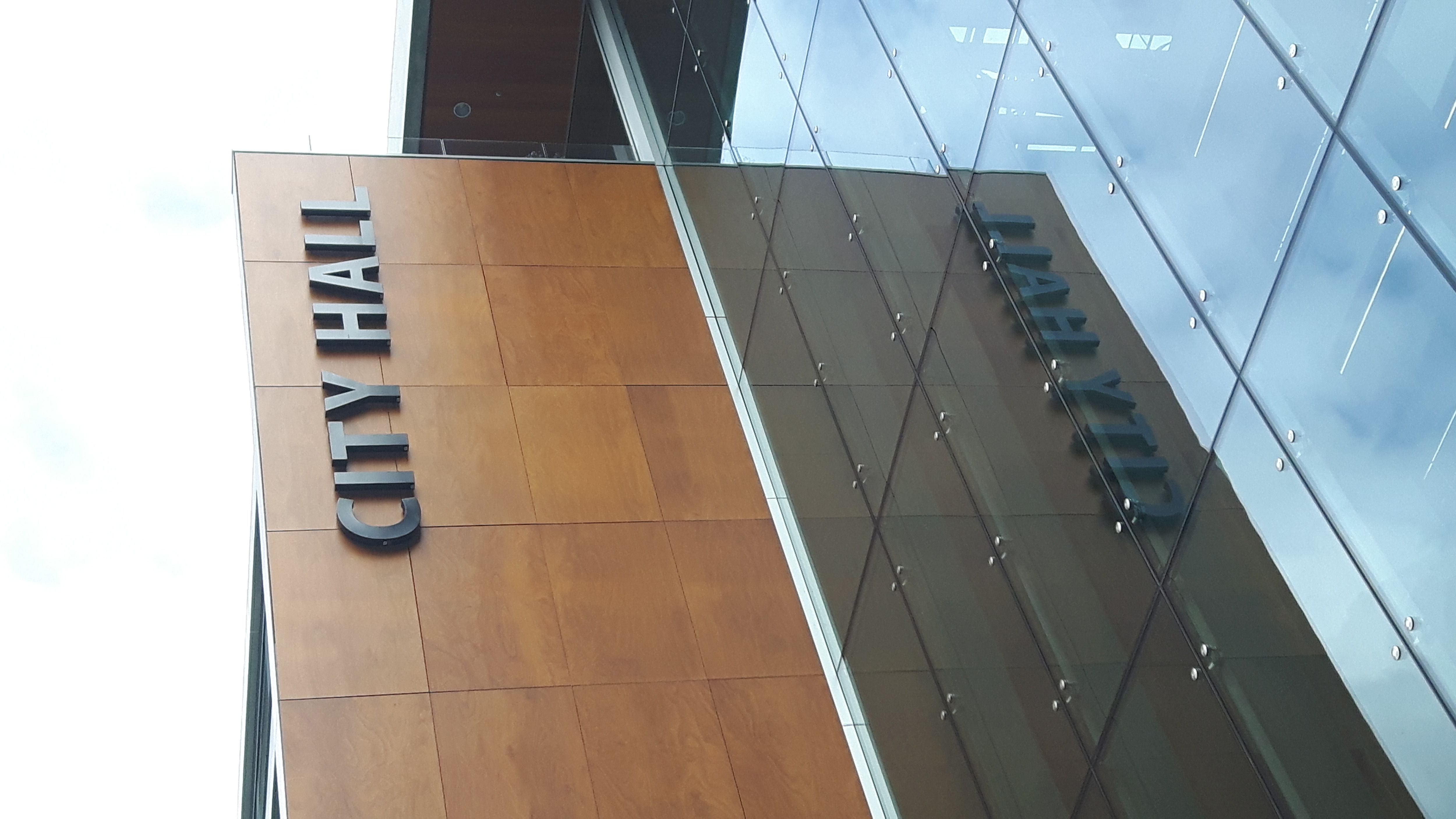A modern building features a striking combination of glass and wood materials. The prominent sign reads "CITY HALL" in bold, dark letters, contrasting against the warm wood paneling. Reflections of the sky and surrounding elements can be seen on the glass facade, enhancing the contemporary aesthetic.