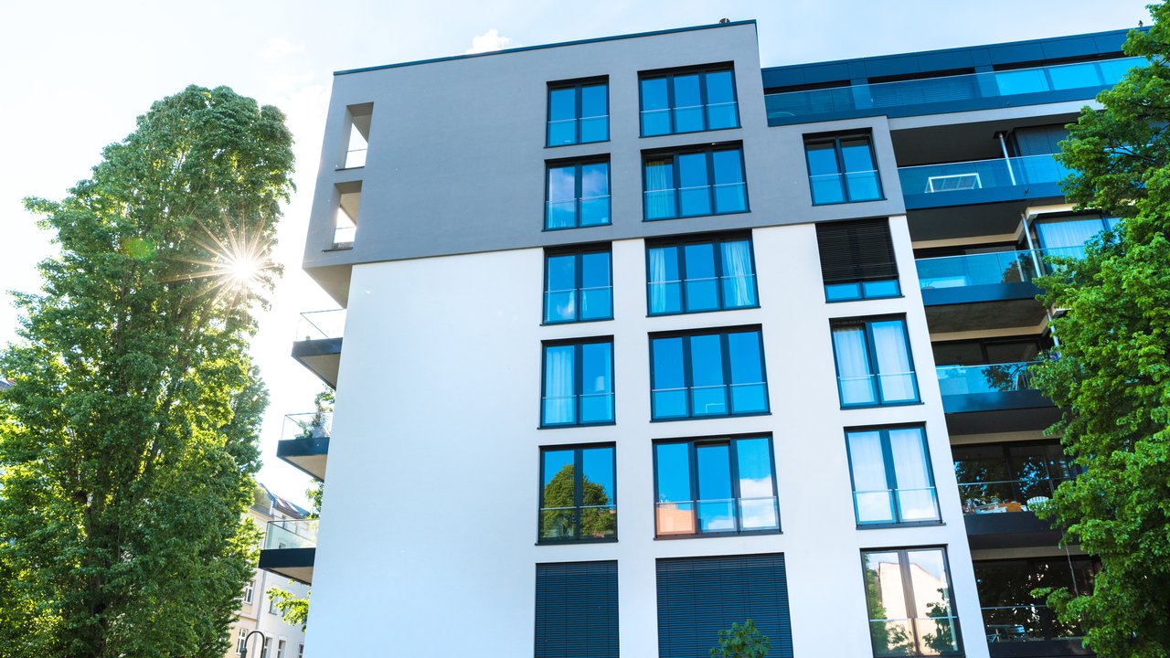 A modern apartment building with large windows and balconies, surrounded by trees and blue sky.