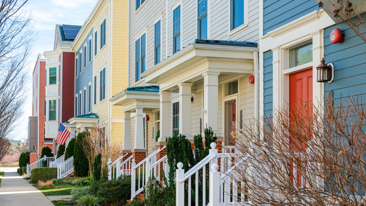 A row of colorful townhouses features a mix of architectural styles, showcasing shades of blue, yellow, and red. Each home has a welcoming porch with white railings and neatly trimmed shrubs. An American flag adds a patriotic touch, while solar panels hint at modern sustainability. The scene is bright and inviting.