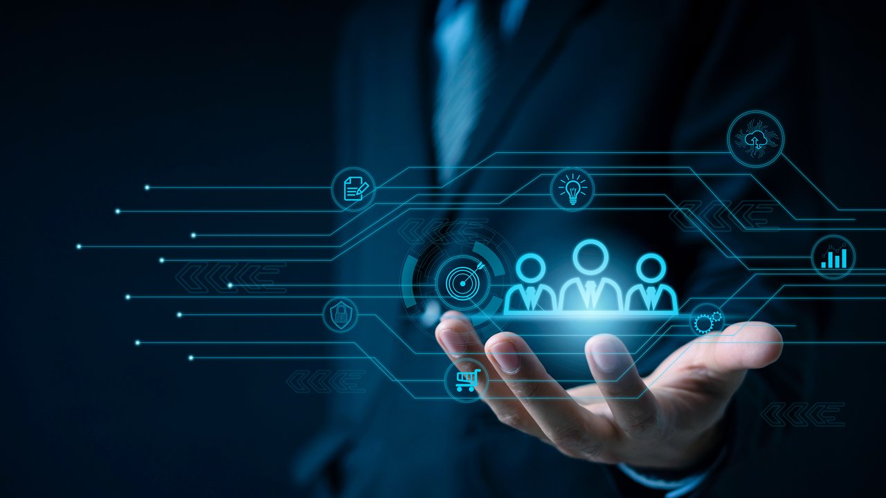  A hand extends forward, palm up, against a dark background. Above it, glowing digital icons represent various concepts like teamwork, innovation, and data analysis. Lines connect these icons, suggesting a network or flow of information. The overall theme conveys technology and collaboration in a professional context.