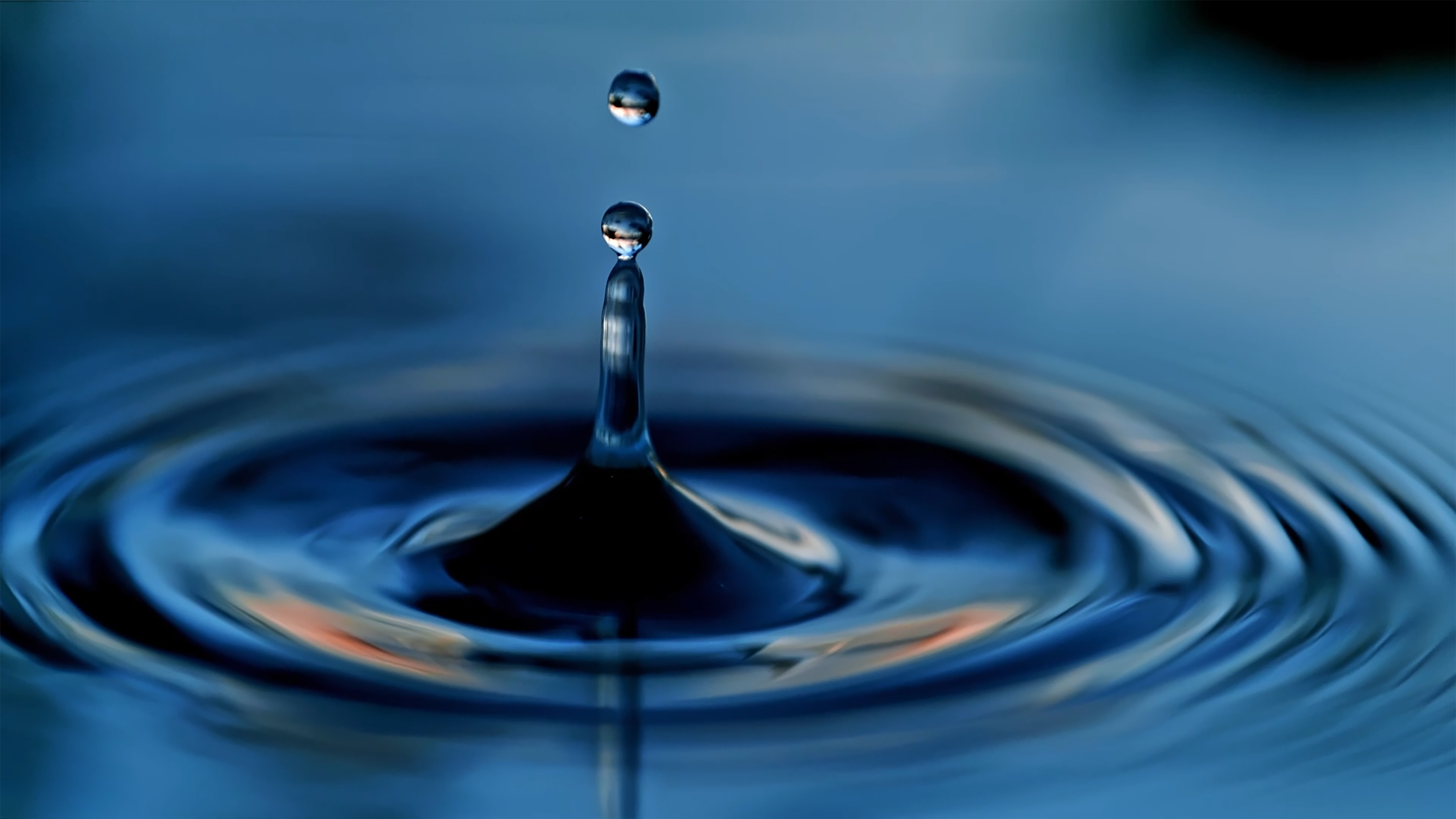 A close-up view captures a droplet of water poised above a circular ripple on a deep blue surface. The droplet reflects light, creating a shimmering effect, while the surrounding ripples radiate outward, enhancing the sense of motion. The smooth textures and rich colors evoke a tranquil, fluid atmosphere.