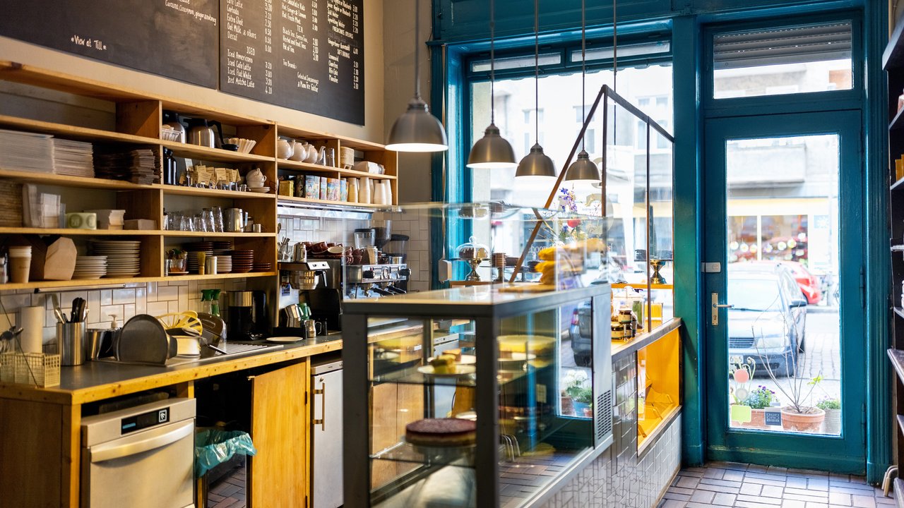 A cozy café interior features wooden shelves filled with dishes and drinks, a sleek counter with a coffee machine, and a glass display case showcasing pastries. Bright blue window frames and pendant lights create a welcoming atmosphere. Outside, colorful cars and plants add to the lively street view.