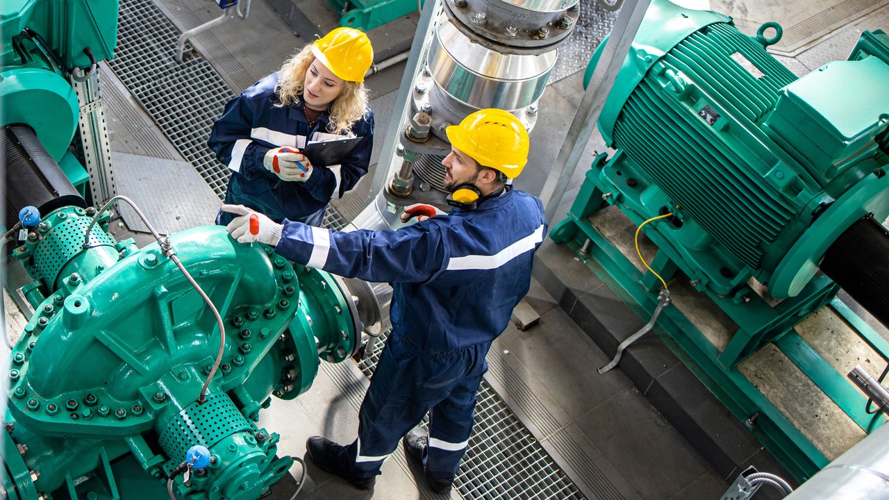 In a mechanical setting, two individuals in safety gear, including helmets and gloves, engage with large, turquoise machinery. One person examines a component while the other holds a clipboard, suggesting a collaborative inspection or maintenance task. The environment features metallic pipes and a textured floor, emphasizing industrial functionality.