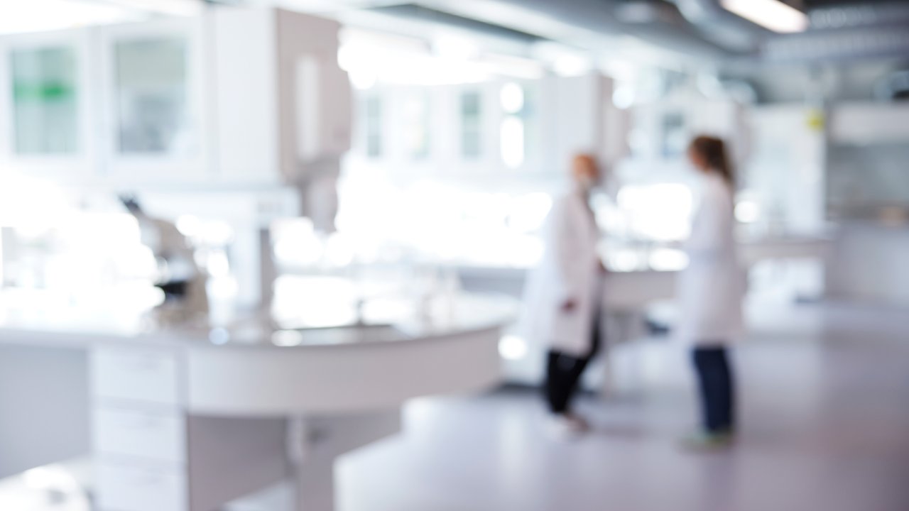 A bright, modern laboratory features sleek, white countertops and equipment. Two figures in lab coats converse in the foreground, with a microscope visible on a nearby table. The background is softly blurred, emphasizing the clean, spacious environment filled with natural light, suggesting a collaborative scientific atmosphere.