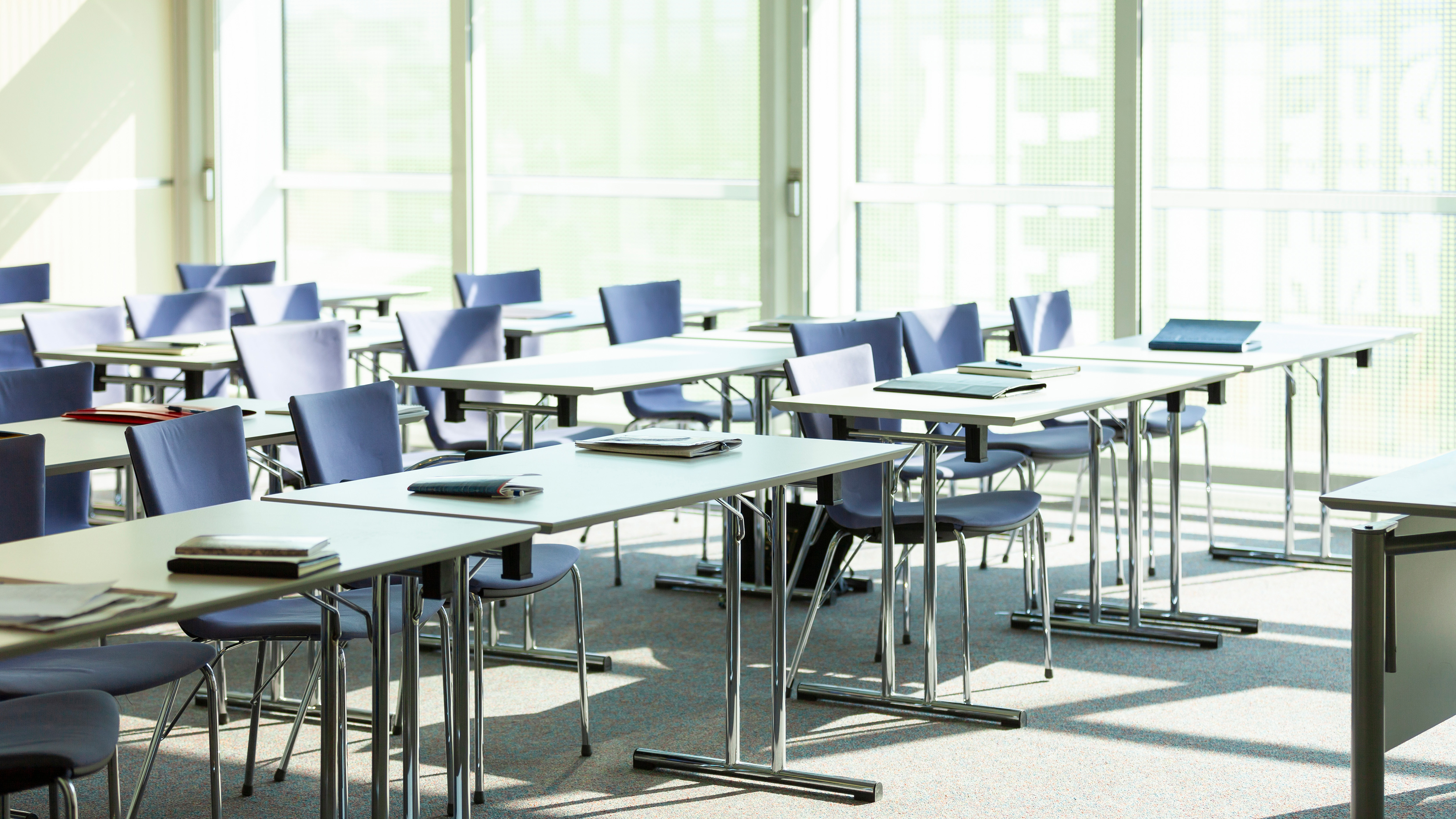 A classroom with multiple tables and chairs, some with books, and large windows allowing natural light.