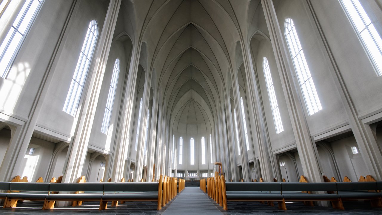 jpg  A spacious interior features soaring, vaulted ceilings with sharp angles, creating a sense of height and openness. Tall, narrow windows allow natural light to flood in, casting soft shadows on the stone floor. Simple wooden pews line the sides, enhancing the minimalist aesthetic of the space.
