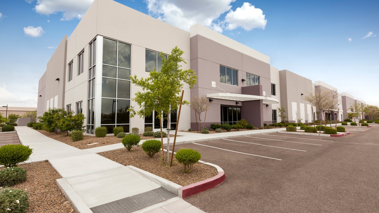  A modern commercial building features a mix of light and dark gray exteriors with large glass windows. Surrounding the structure are neatly trimmed shrubs and small trees, adding greenery to the landscape. A wide, accessible walkway leads to the entrance, while a spacious parking lot is visible in the foreground.