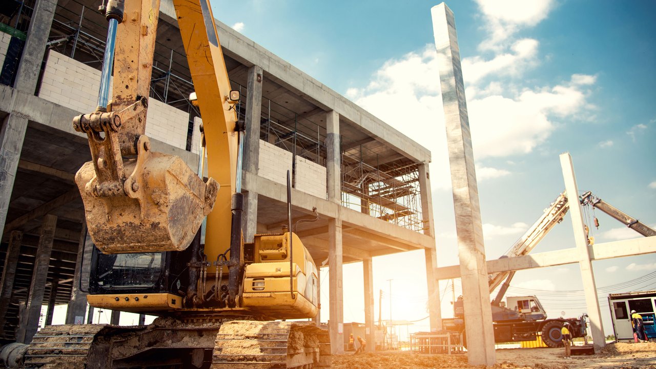 A construction site features a large yellow excavator with a dirty bucket, positioned prominently in the foreground. Behind it, a partially built concrete structure rises, showcasing scaffolding and unfinished walls. The sky is bright with scattered clouds, and sunlight casts a warm glow, enhancing the scene's industrious atmosphere.
