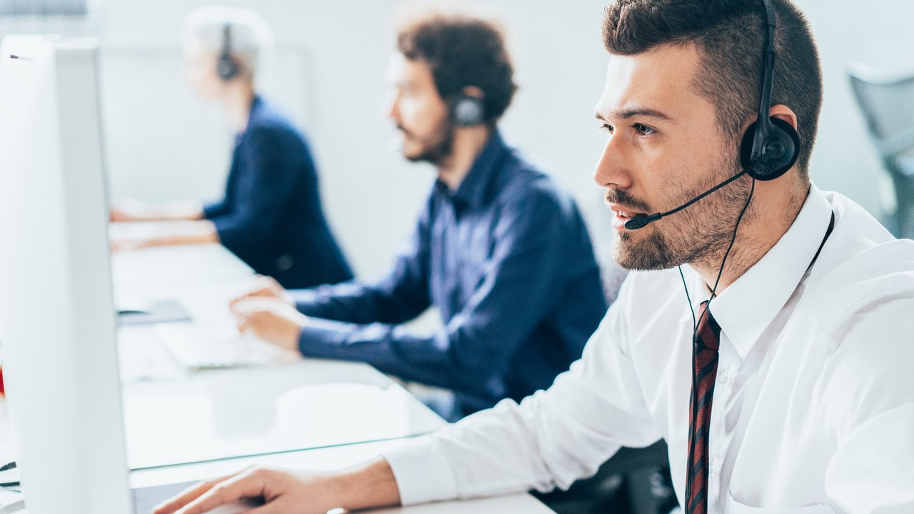 Three people wearing headsets work at computers in a bright office setting.