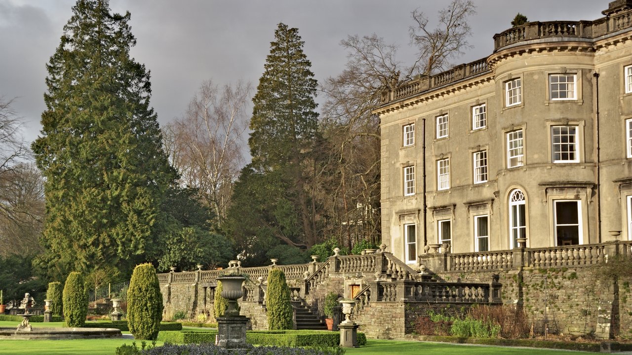 A large stone mansion with a landscaped garden, featuring trees, shrubs, and ornamental urns.