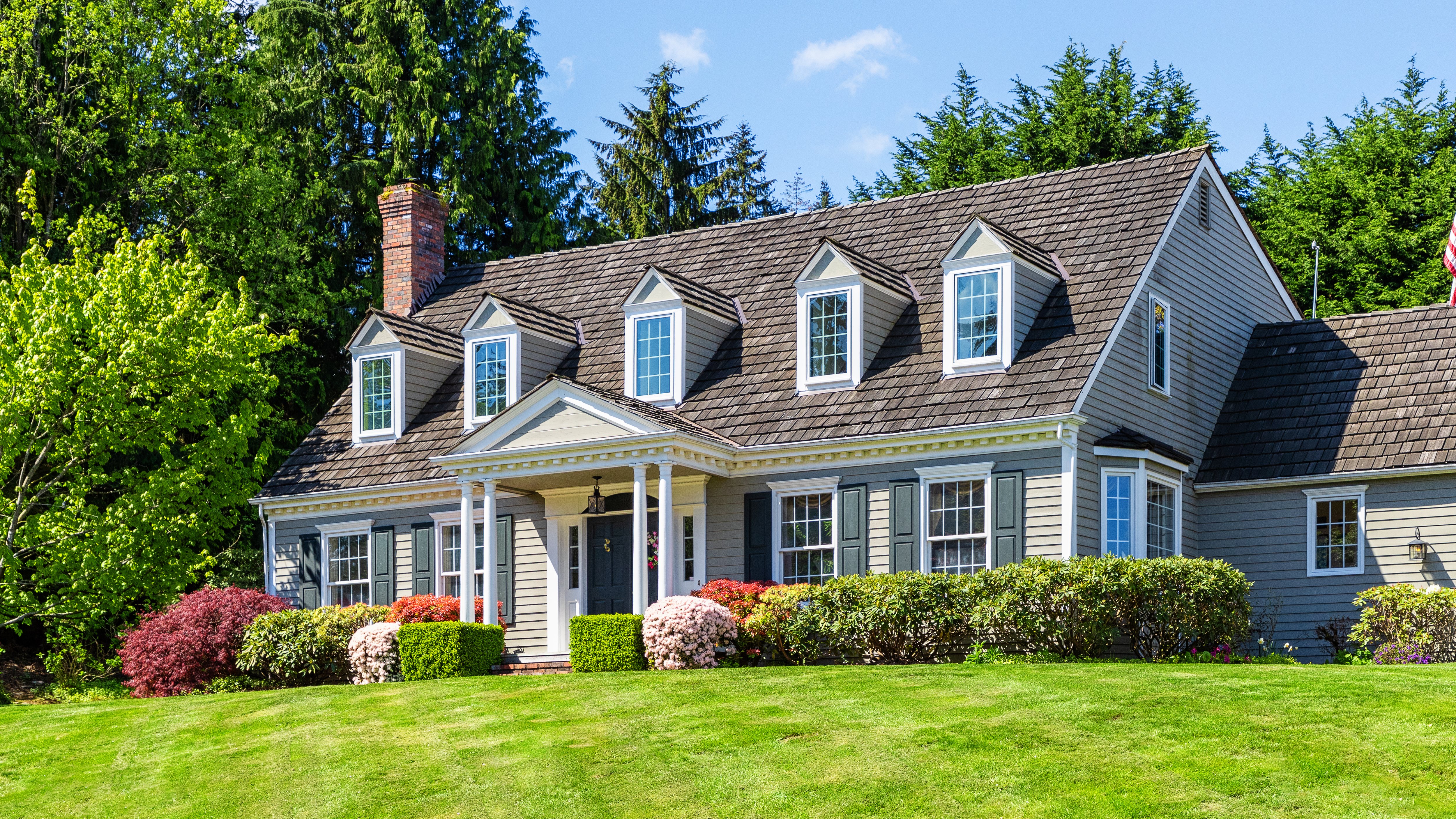 A large, two-story house with a sloped roof, surrounded by green grass and colorful shrubs under a clear blue sky.