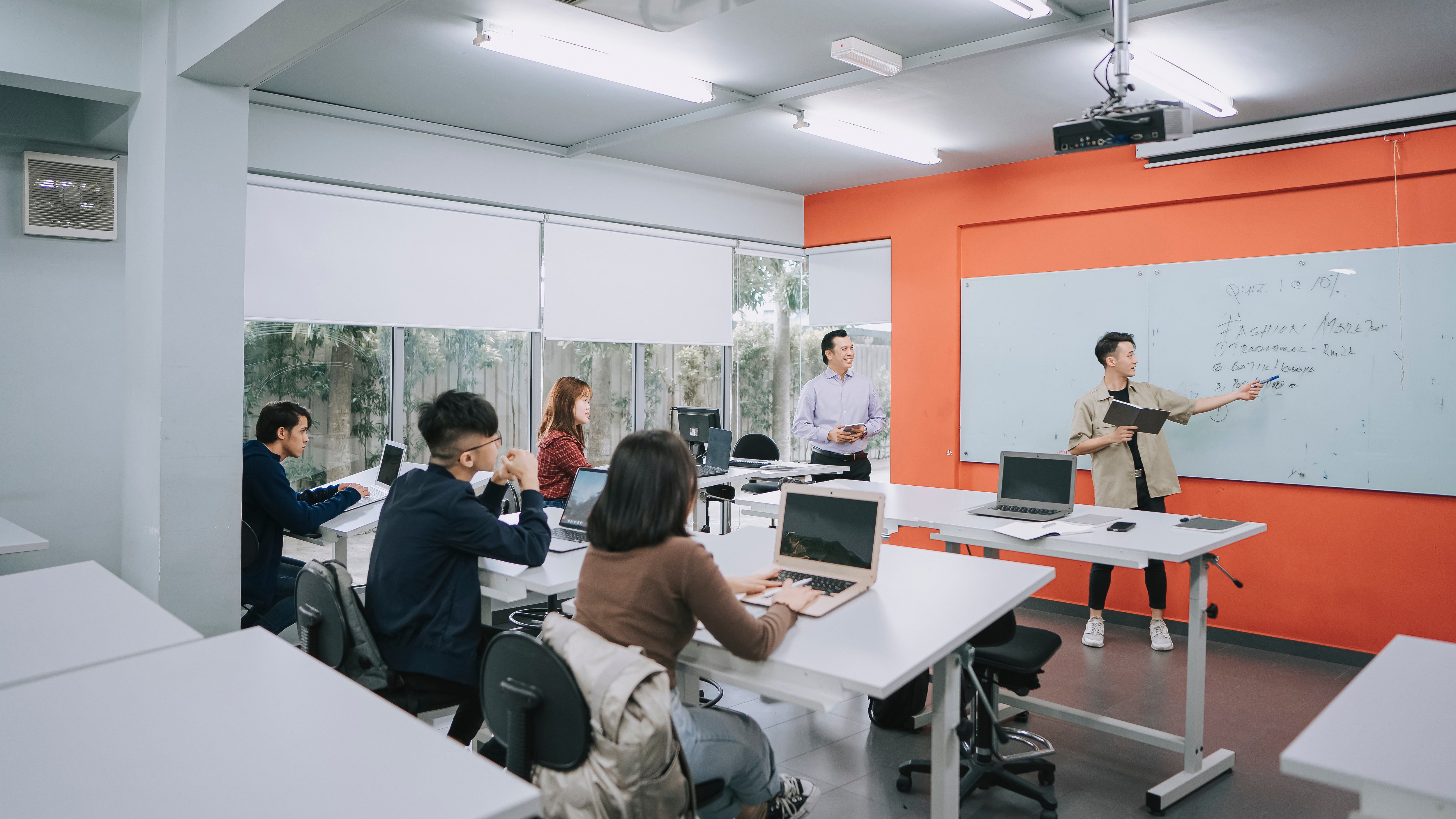 A classroom with students at desks, a teacher presenting at a whiteboard, and laptops on tables.