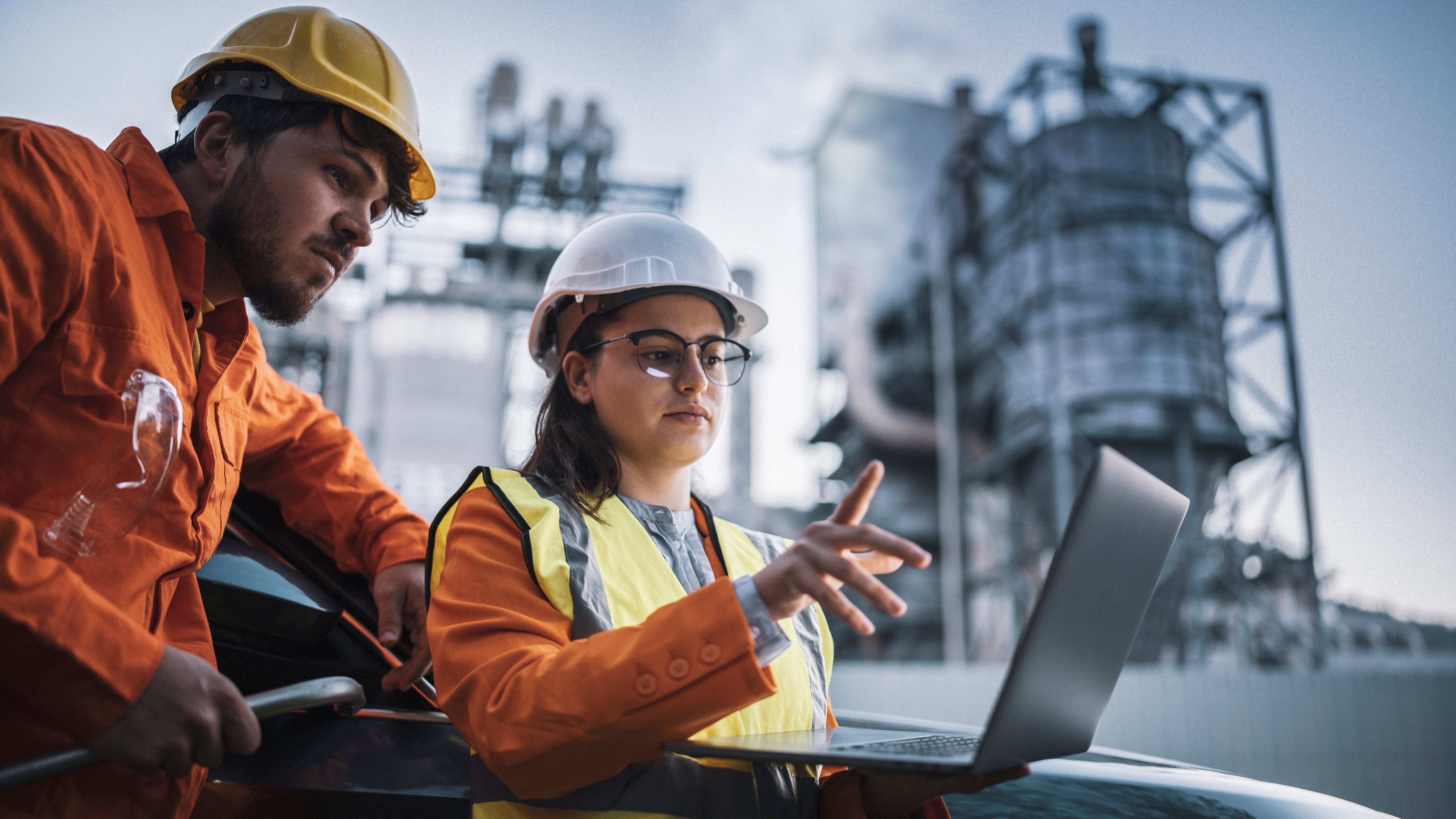 Two workers in safety gear discuss over a laptop near industrial buildings.