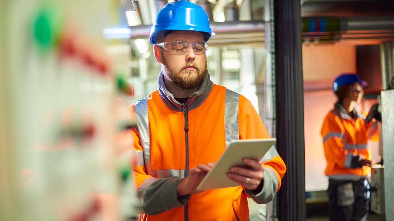 ASME Inspection Services An industrial service engineer conducts a safety check of a control panel in a boiler room. He is wearing hi vis, hard hat, safety glasses and holding a digital tablet as he conducts a safety inspection.