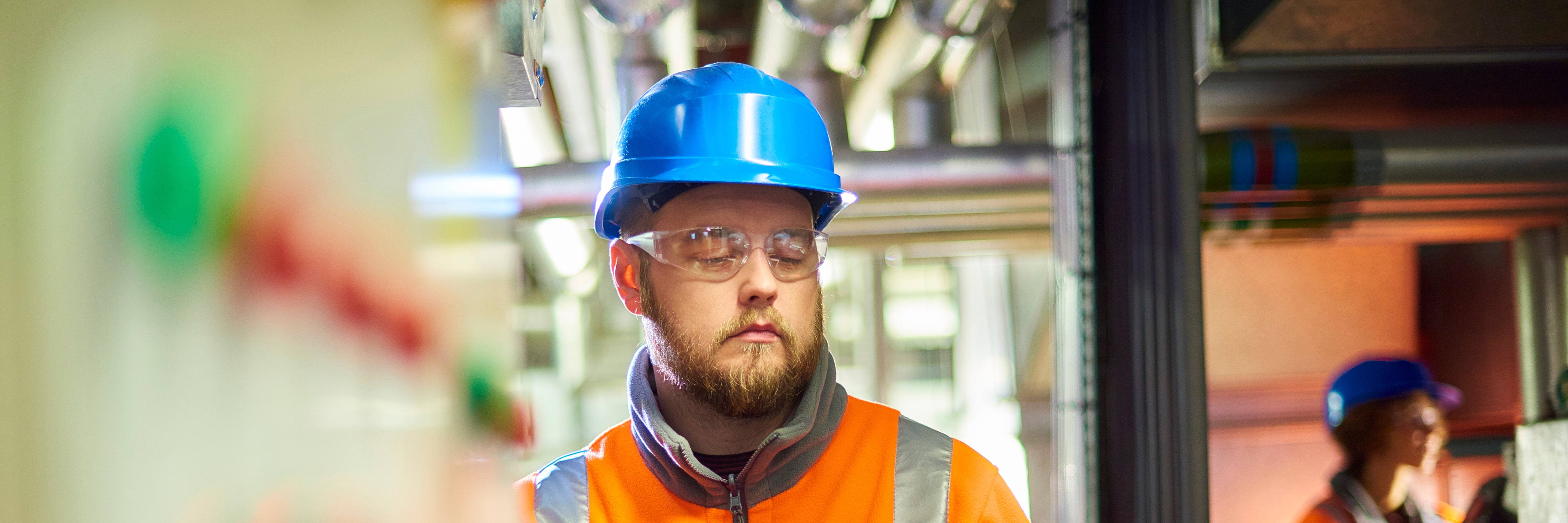 An industrial service engineer conducts a safety check of a control panel in a boiler room. He is wearing hi vis, hard hat, safety glasses and holding a digital tablet as he conducts a safety inspection.