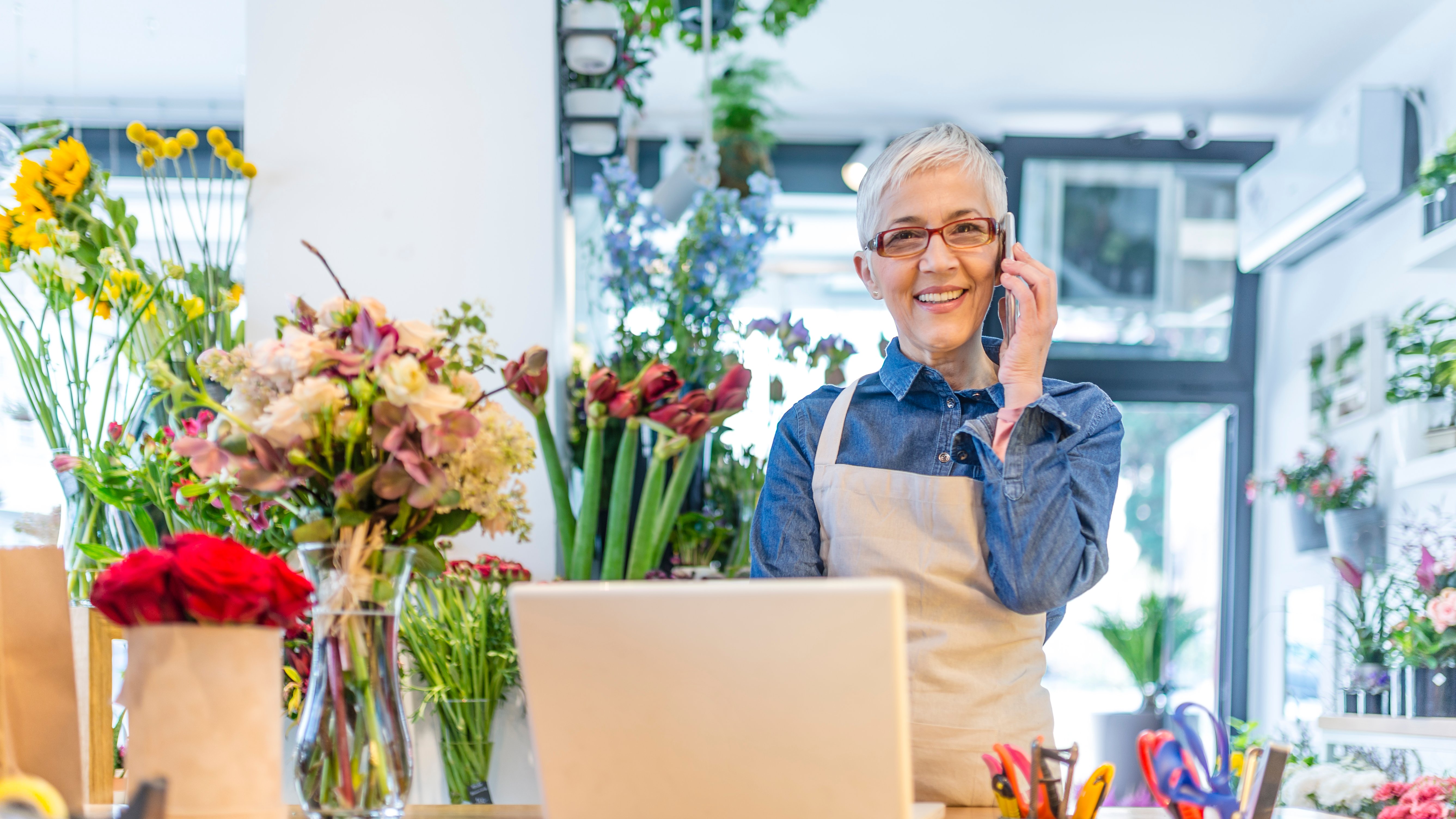 Mature, gray hair, Flower shop worker using a smartphone and a laptop computer in her workplace and smiling while wearing denim shirt and apron. People, business, sale and floristry concept. Photo of Smiling female florist taking order on mobile phone at flower shop.