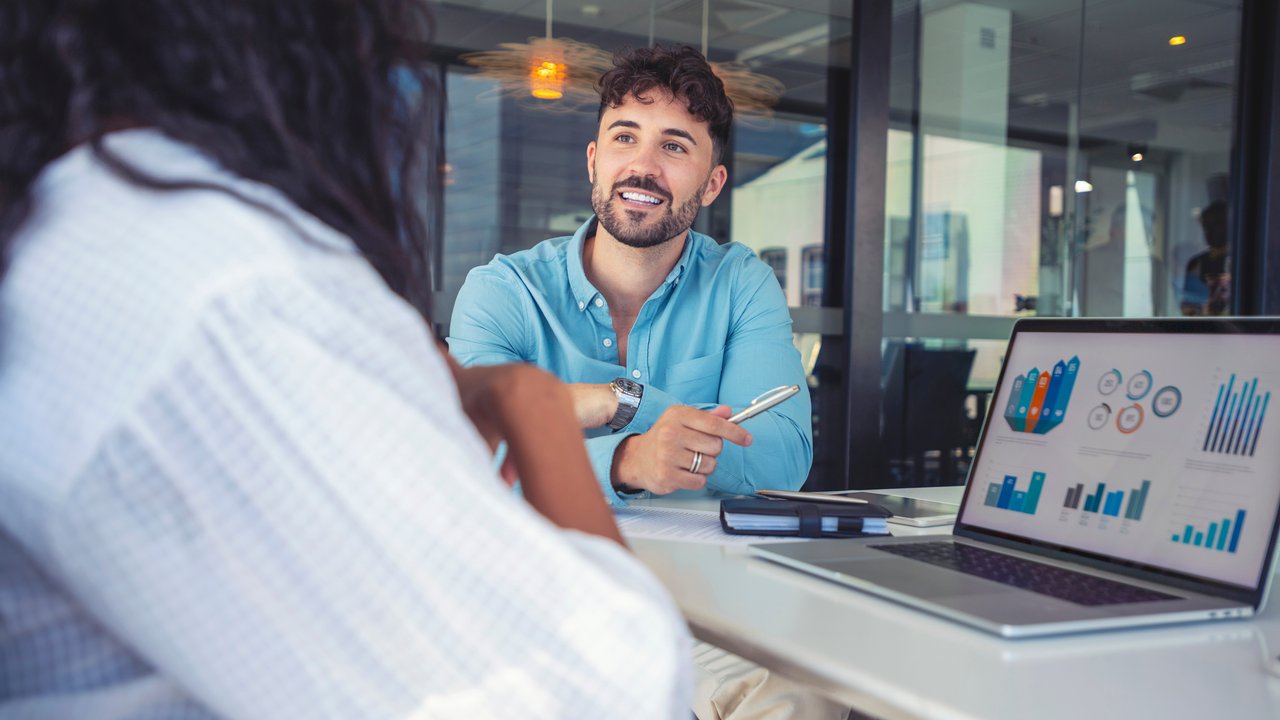 Young professional man discussing charts on laptop in office. Business colleagues having a conversation. They are discussing a charts and graphs on a laptop computer. They are both young business people casually dressed in an office. Could be an interview or consultant working with a client.