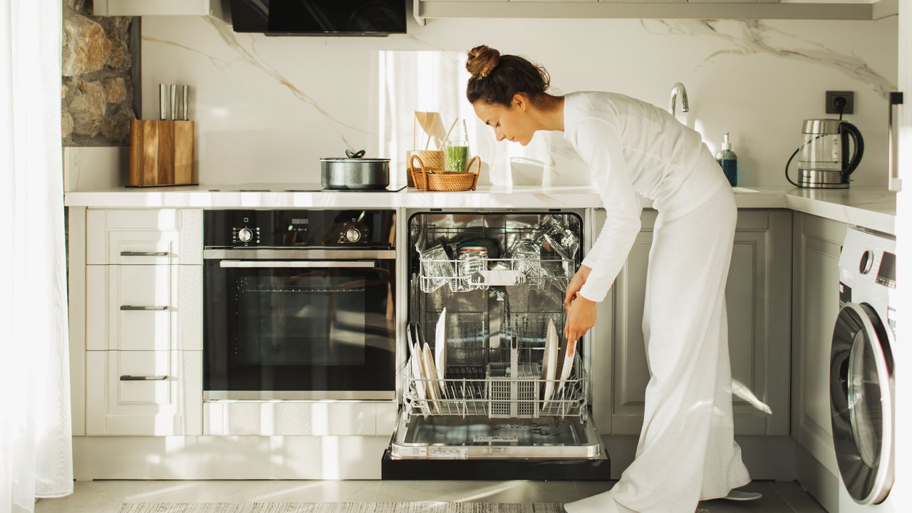 A woman in white attire is loading a dishwasher in a modern kitchen with a stone wall and various appliances.