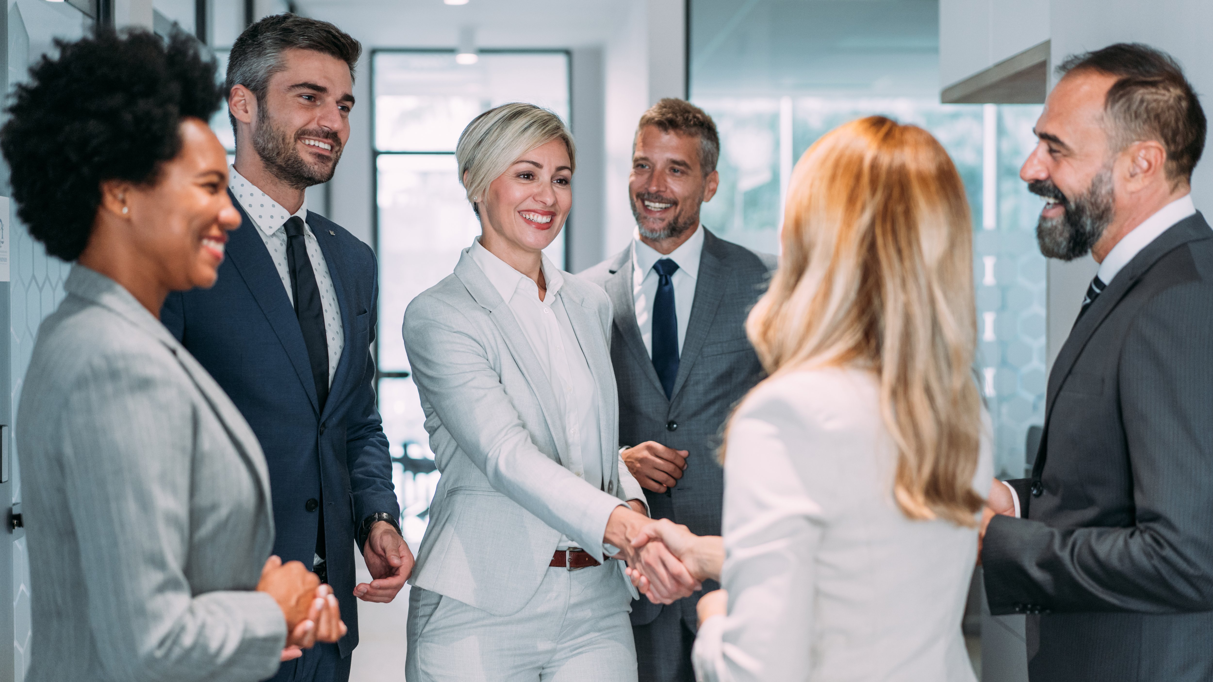 A group of six individuals stands in a modern office setting, engaged in a handshake. They are dressed in professional attire, with a mix of light and dark suits. The background features large windows allowing natural light, enhancing the contemporary atmosphere. The scene conveys a sense of collaboration and professionalism.