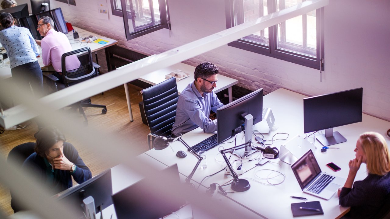 A modern office space features several workstations with computers and monitors. Natural light streams through large windows, illuminating the wooden floor. Individuals are engaged in various tasks, with some collaborating at a desk. The atmosphere is dynamic and focused, reflecting a productive work environment.
