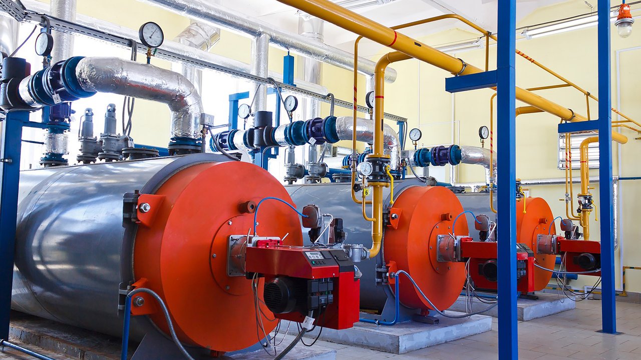 Three large industrial boilers are arranged in a row, each with a prominent orange front and a metallic body. Colorful pipes, primarily yellow and blue, connect them to a network of gauges and valves above. The bright, well-lit room features a clean, tiled floor, emphasizing the organized layout of the machinery.