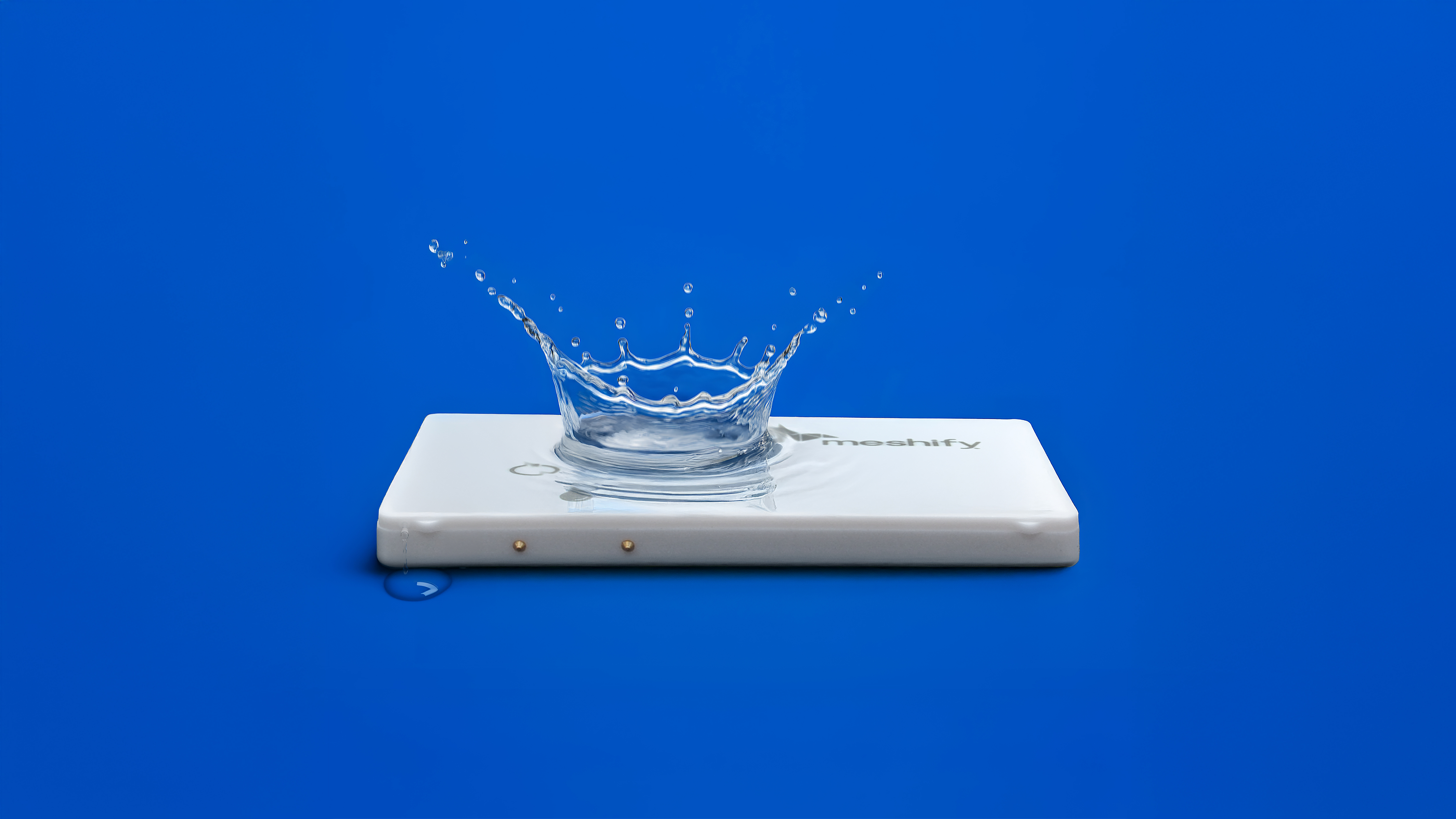 A white rectangular device rests on a vibrant blue background, with a splash of water creating a crown shape above it. The splash features multiple droplets, emphasizing motion and fluidity. The device has a logo and two small indicator lights, adding a modern touch to the scene.