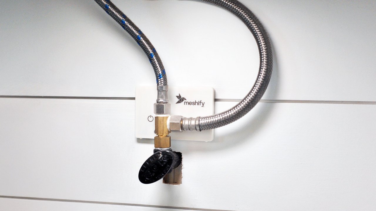 A metal valve with a black handle is mounted on a wall, next to a white control panel featuring a power symbol. A flexible, braided hose connects the valve to the panel, which is branded with the word "meshify." The background consists of light-colored wooden planks, creating a clean, modern aesthetic.