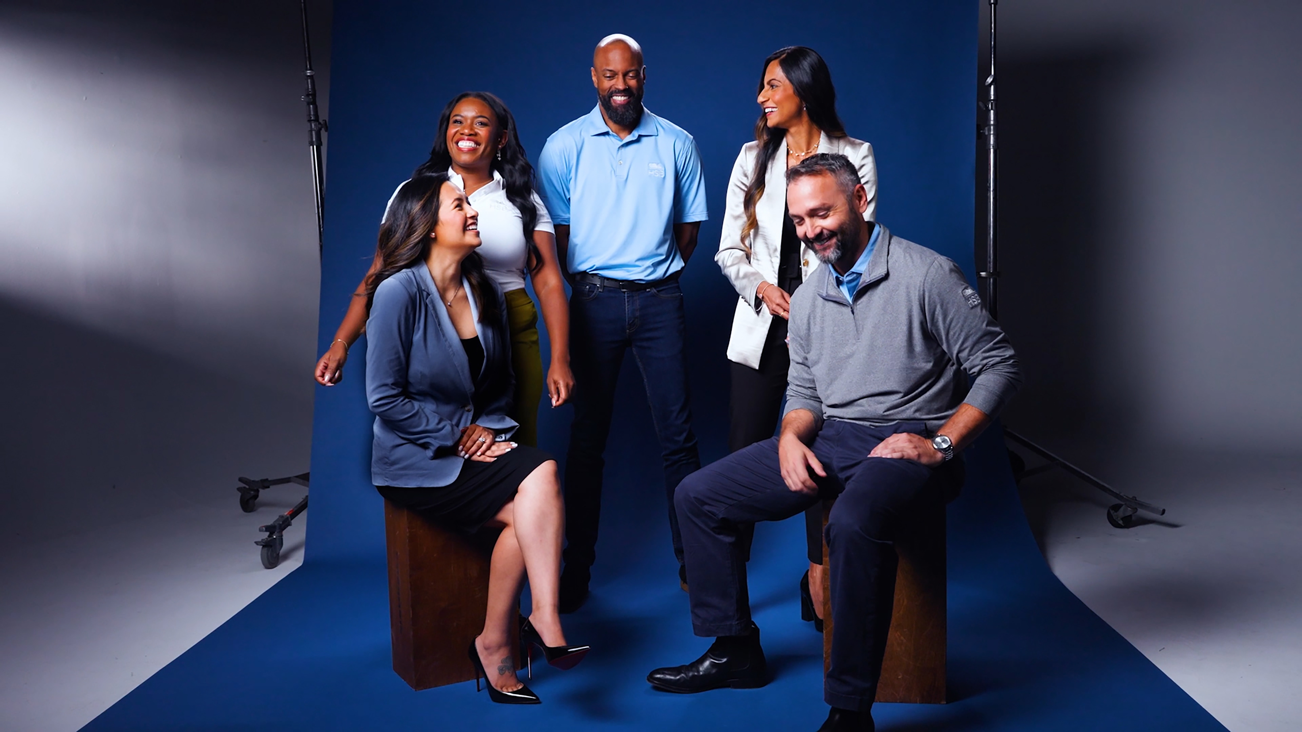 A group of five individuals poses in a studio setting with a deep blue backdrop. They are dressed in a mix of professional and casual attire, showcasing a variety of styles. The composition features two seated figures on wooden blocks, while the others stand behind them, creating a dynamic arrangement. Soft lighting highlights their expressions.