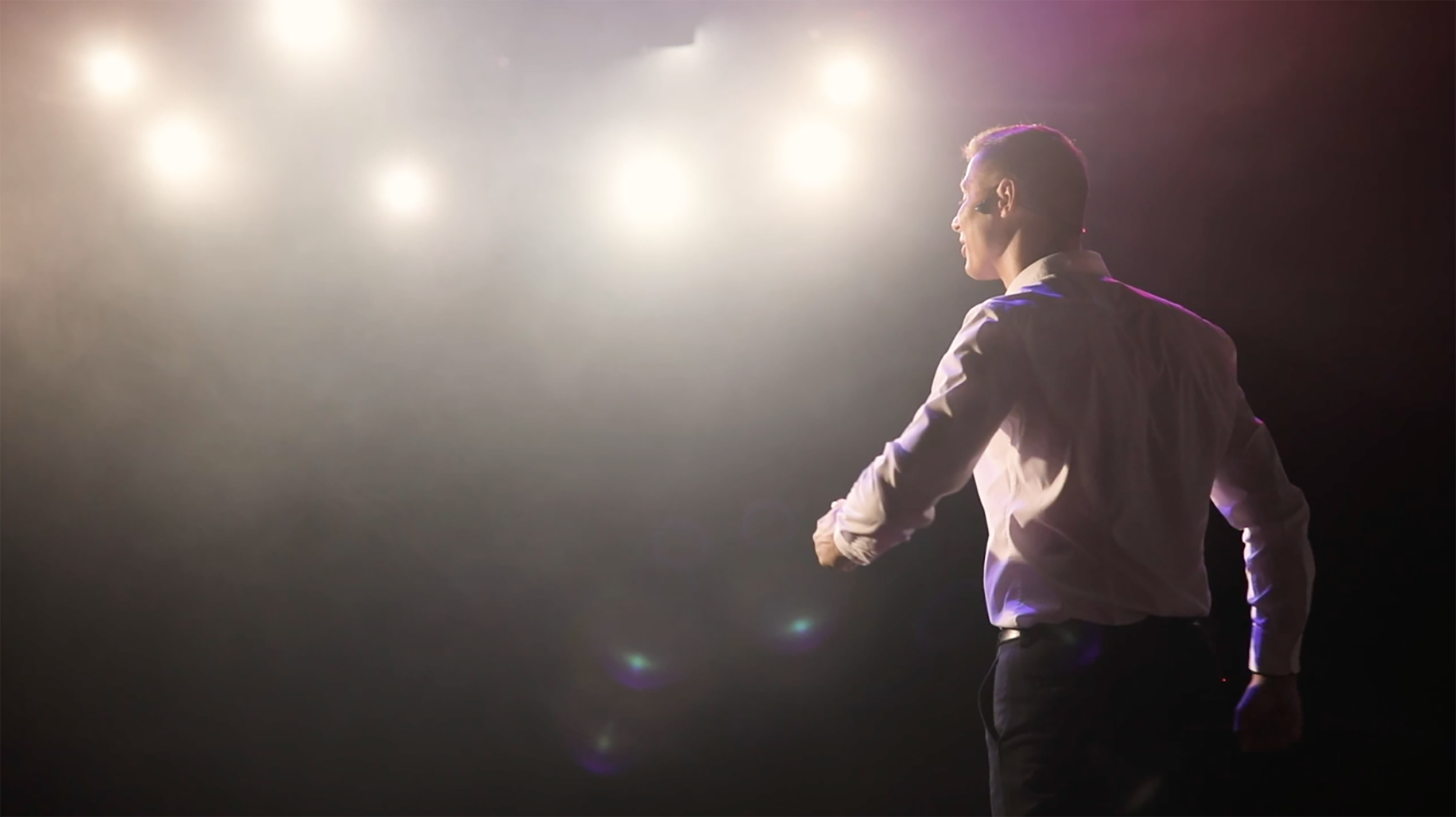 Man standing on stage facing audience under lights.