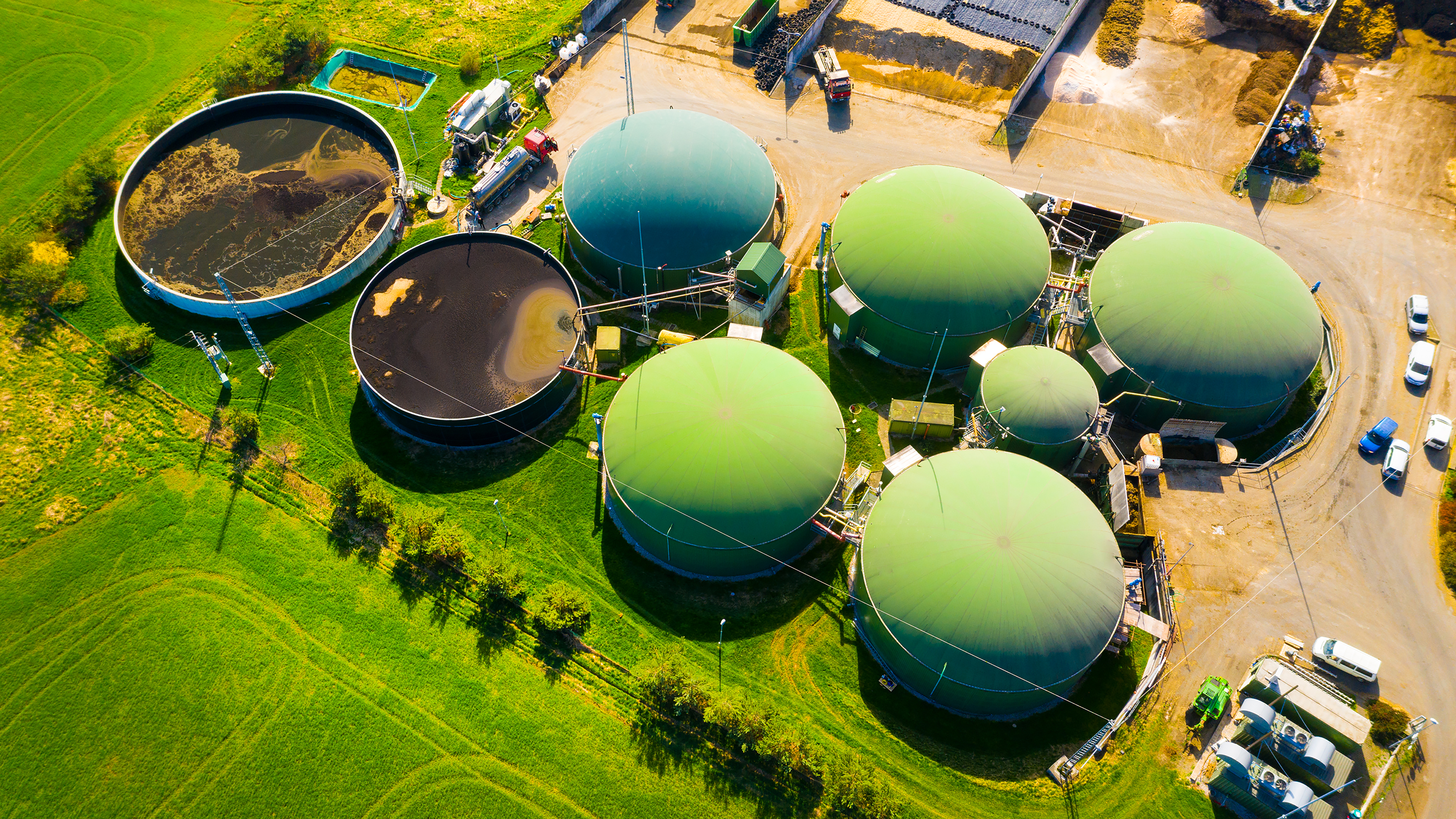 Aerial view of a waste treatment facility featuring several large, green domes and circular tanks. The domes are connected by pipes, with some tanks containing dark liquid. Surrounding the facility are lush green fields and a dirt road with parked vehicles. The layout emphasizes the integration of technology and nature in waste management.