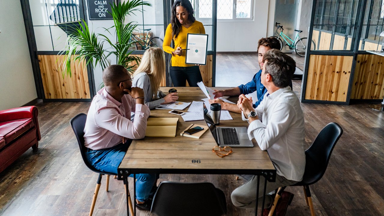 HSB Expands Cyber Coverage A diverse group of individuals is engaged in a meeting around a wooden table in a modern office space. One person stands, presenting a document, while others are seated, reviewing papers and a laptop. The room features large windows, plants, and a motivational poster, creating an inviting atmosphere.
