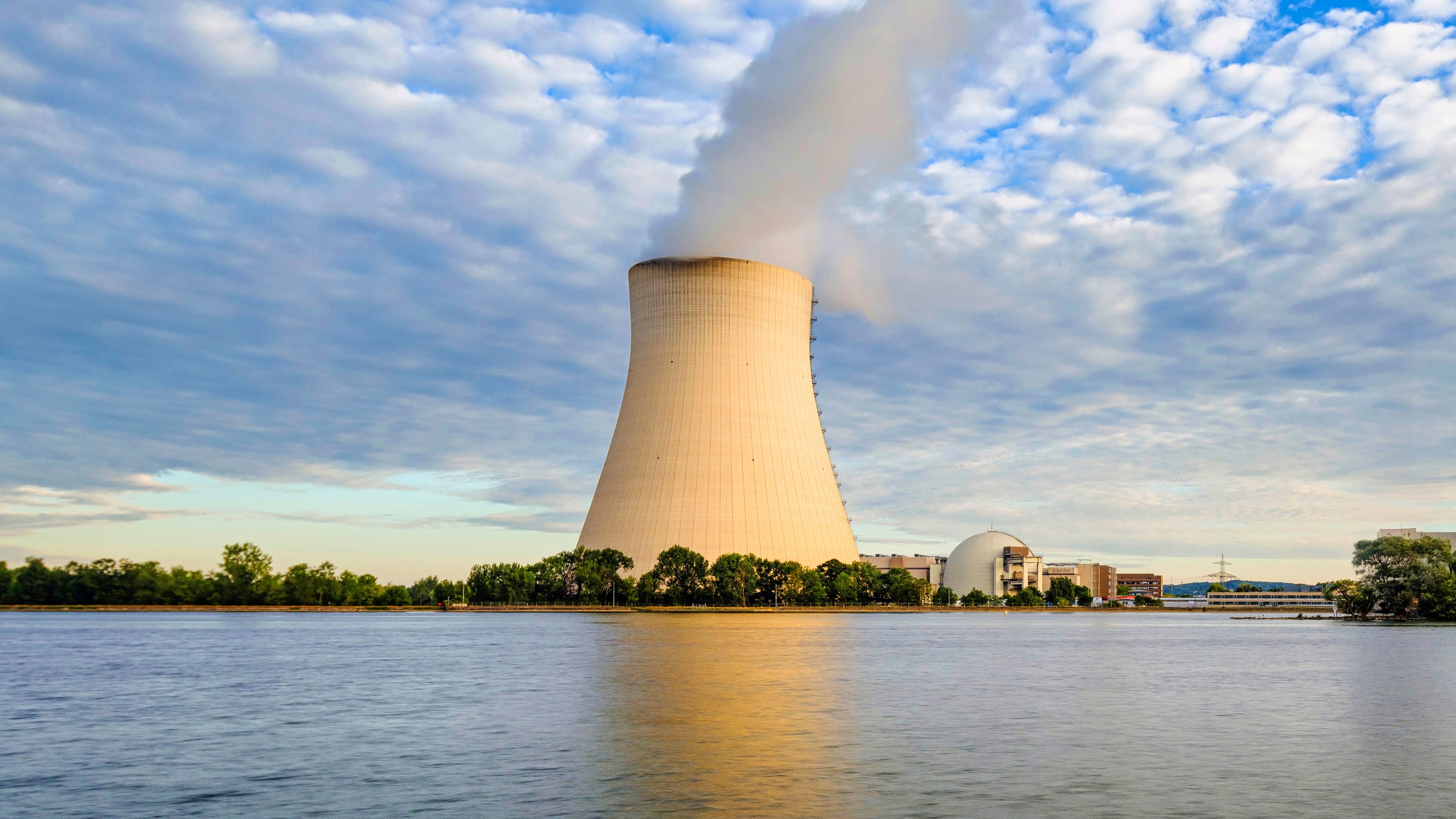 Nuclear Services A cooling tower emits steam near a river, with a power plant and trees in the background under a cloudy sky.
