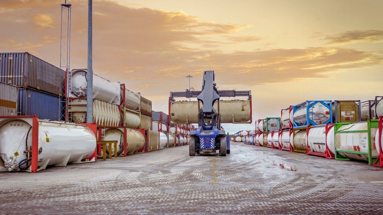 A large blue forklift is positioned centrally, lifting a white cylindrical container above the ground. Surrounding it are rows of stacked containers in various colors, including green, red, and blue, creating a vibrant industrial scene. The sky features soft orange and purple hues, indicating sunrise or sunset, enhancing the atmosphere.