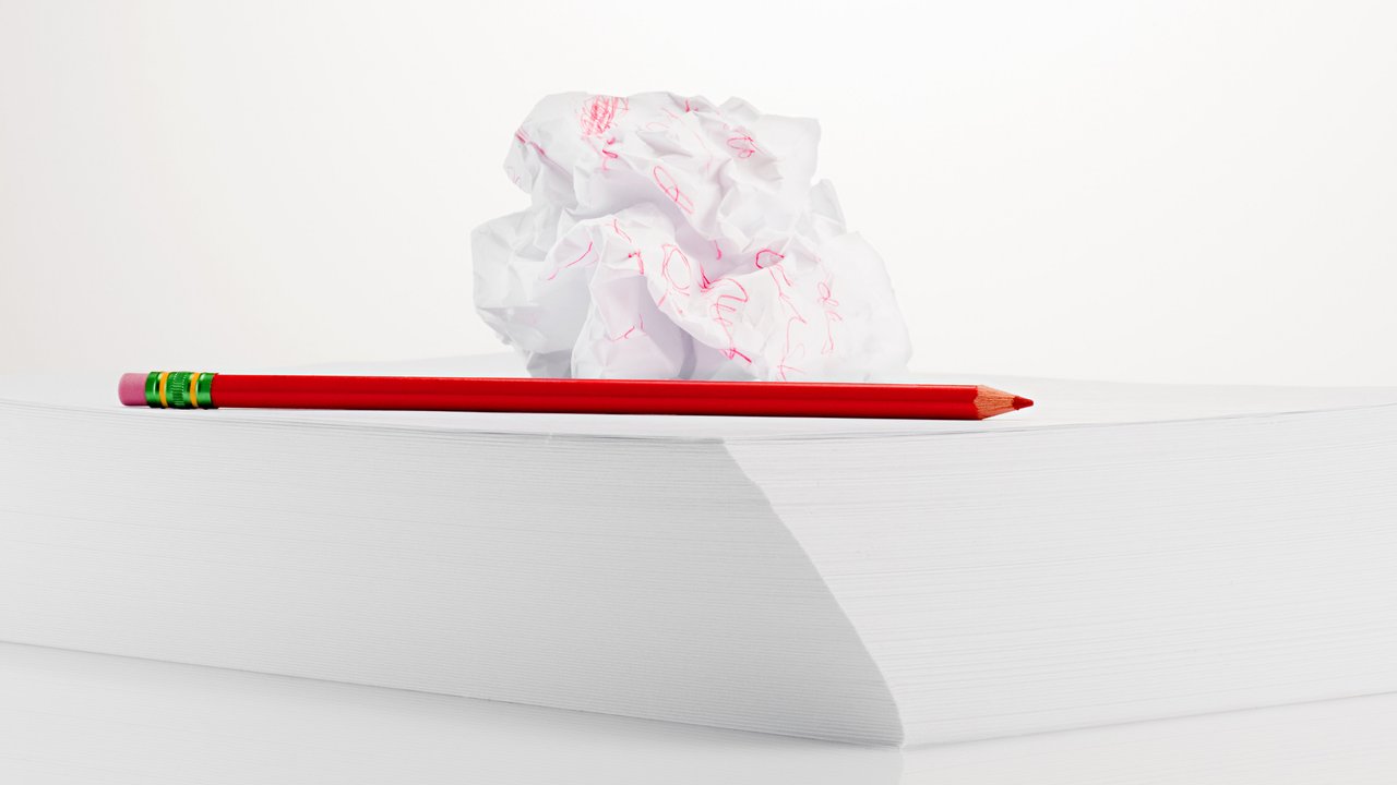 A stack of blank white paper sits on a reflective surface, creating a clean and minimalistic backdrop. A bright red pencil rests alongside the paper, its sharpened tip pointing outward. Above the stack, a crumpled piece of white paper, marked with pink scribbles, adds a touch of creativity and imperfection to the scene.