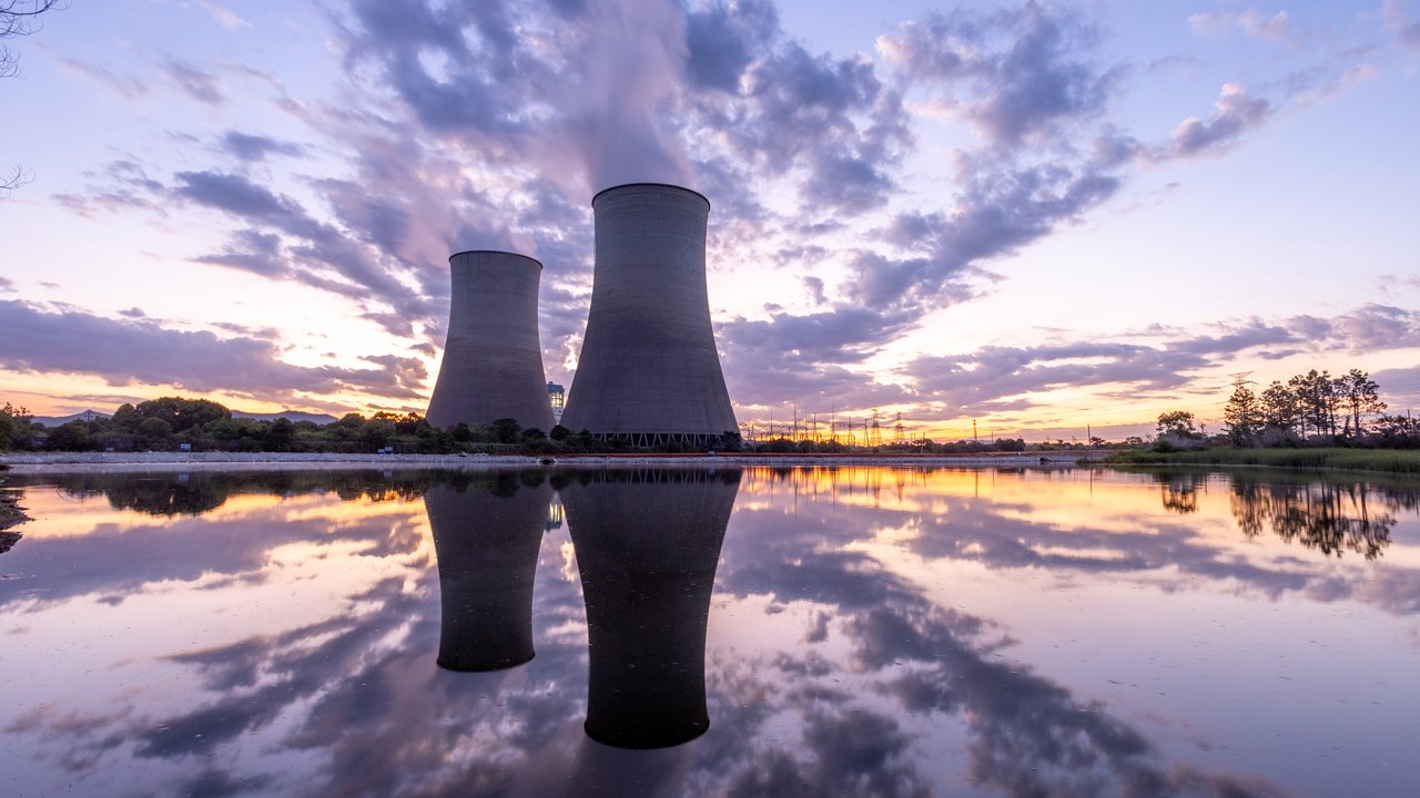 Two large cooling towers rise against a vibrant sunset, surrounded by a tranquil body of water that reflects the scene. Wispy clouds stretch across the sky, blending shades of orange, pink, and blue. The stillness of the water enhances the dramatic visual contrast between the industrial structures and the natural landscape.