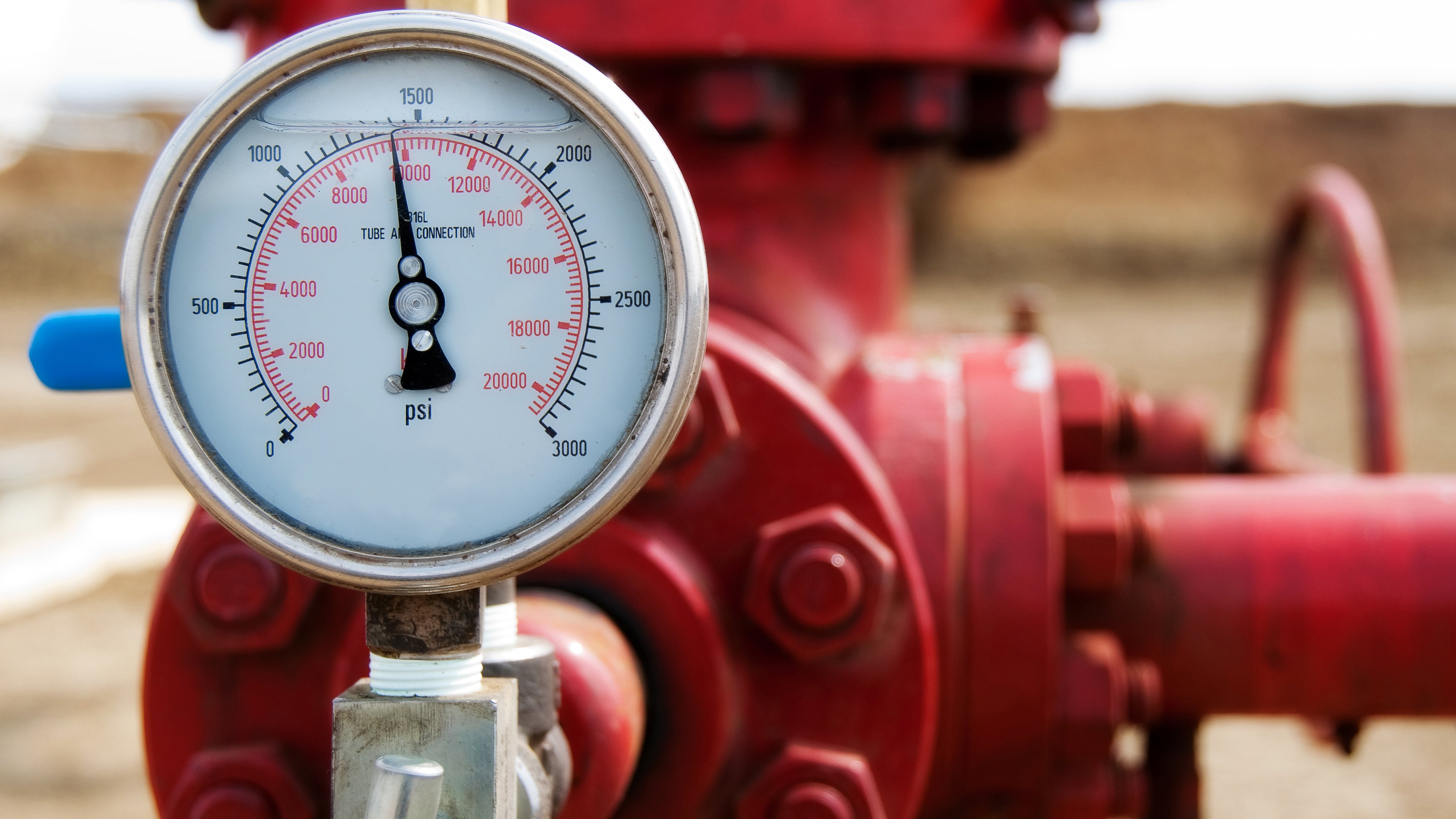 A close-up of a pressure gauge showing psi readings, attached to a red valve and pipe in an outdoor setting.
