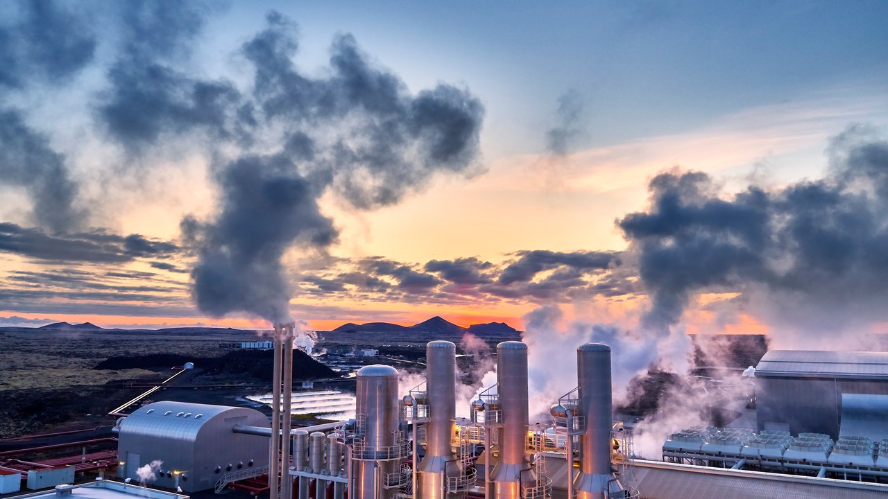 Industrial facility with smokestacks emitting steam against a sunset backdrop and mountains in the distance.
